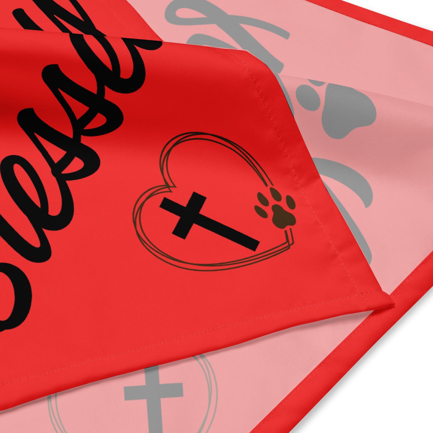 A folded red bandana shows both the front and back of it and the hem. You can see part of the design that is printed on it. You can see part of words and a heart with a cross inside it and a pawprint makes up part of the side of the heart.