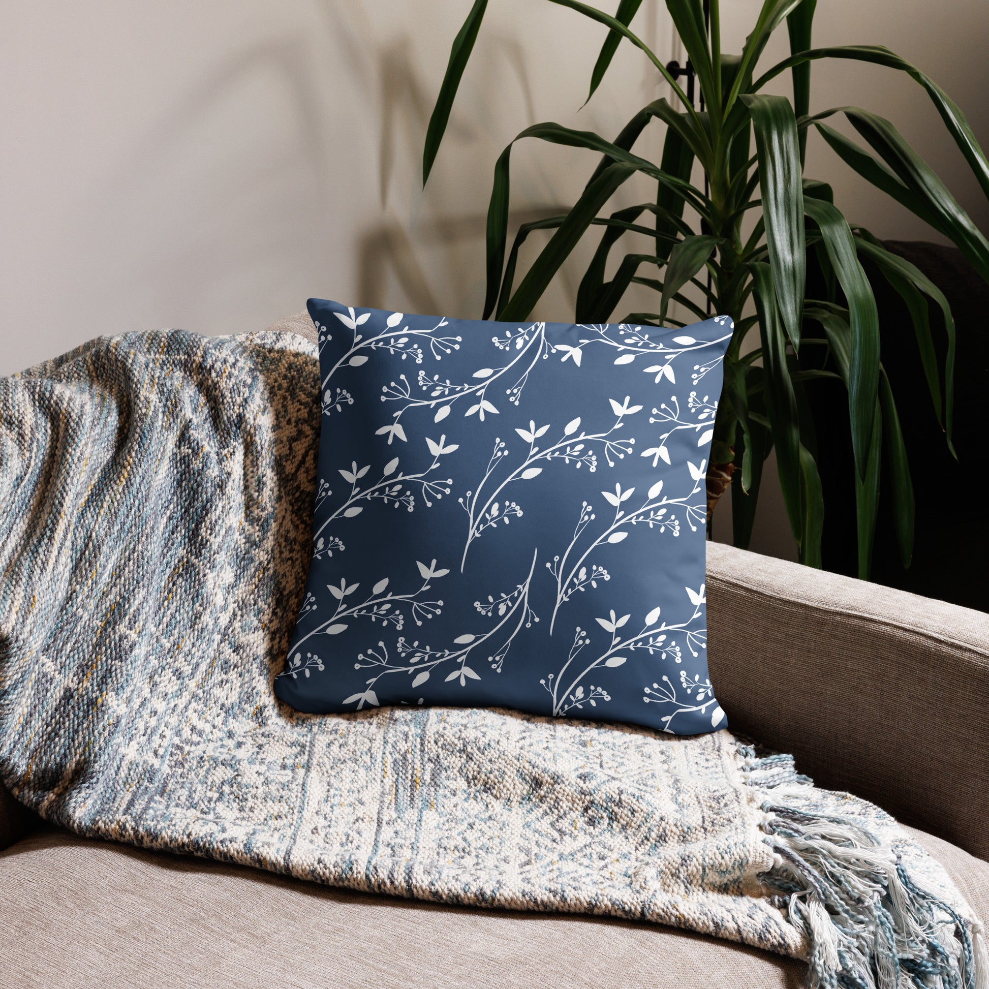 Tan couch with neutral throw with fringes laying across it and the back, and a blue pillow shows its back side which is blue with kind of a vining white flowers on it, and there is a plant behind the couch.