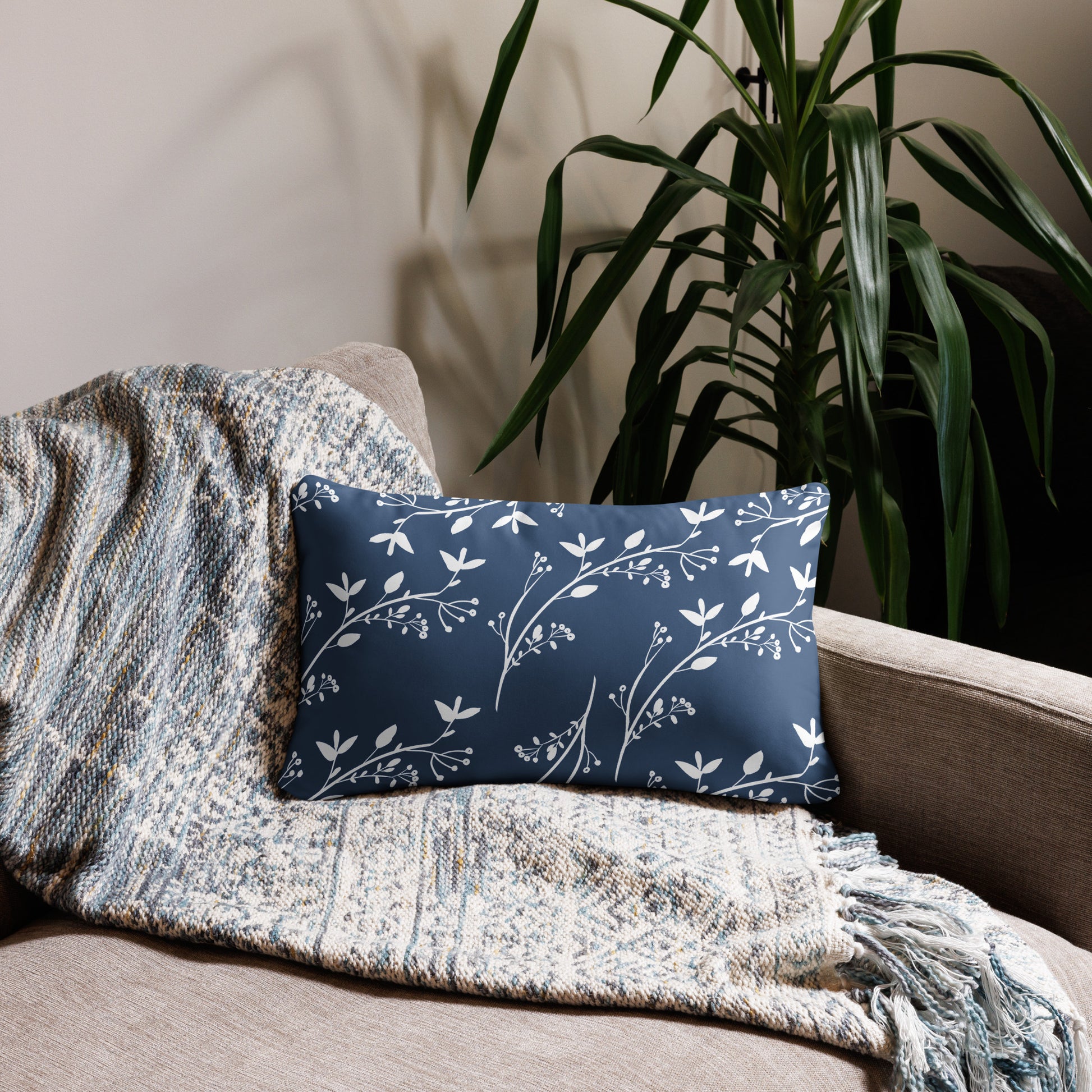 In the middle of the photo, there is a horizontal oblong pillow showing the backside which is blue with white vining flowers all over it. It sits on a couch with a neutral throw, beside a tall green plant behind the couch.