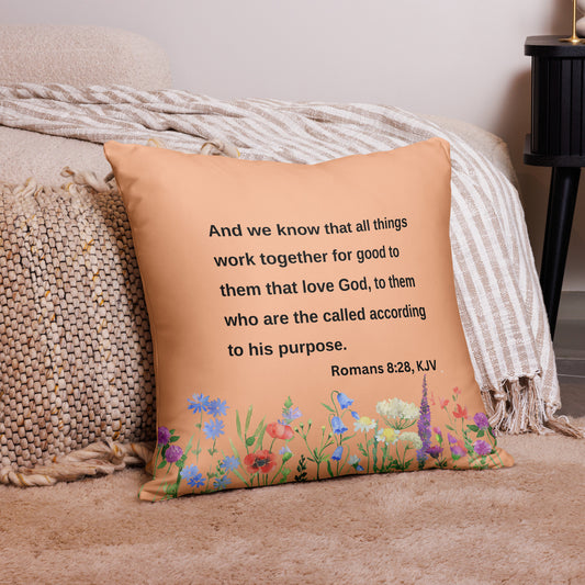 Peach colored pillow with multi-colored wildflowers on the bottom and words in black that say "And we know that all things work together for good to them that love God, to them who are the called according to his purpose. Romans 8:28, KJV" leaning against a bed sitting on carpet.