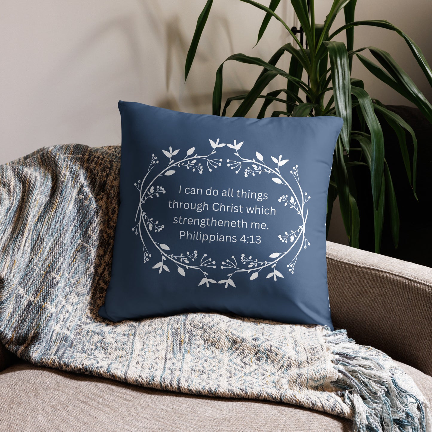 Corner of a light brown neutral couch with a neutral tannish women throw on it and a blue accent or throw pillow in the corner with a white floral wreath printed on it and inside in white text it says, "I can do all things through Christ which strengtheneth me. Philippians 4:13" and there is a green plant behind the couch.