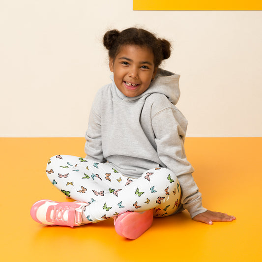 Little girl with dark hair in two buns sitting on yellow flooring cross legged and smiling. She is wearing a light gray sweatshirt, white leggings with a pattern of pink, blue, yellow, and green butterflies, and white and pink shoes.