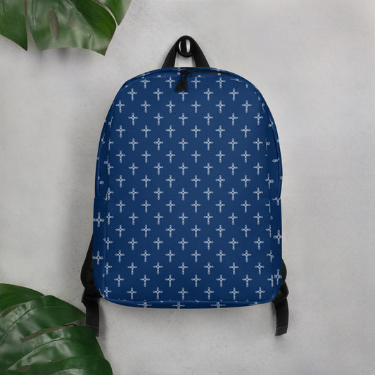 Navy blue backpack with a white ornate cross print and black trim hanging on a hook on a white wall with a few large green leaves in two corners of the photo.