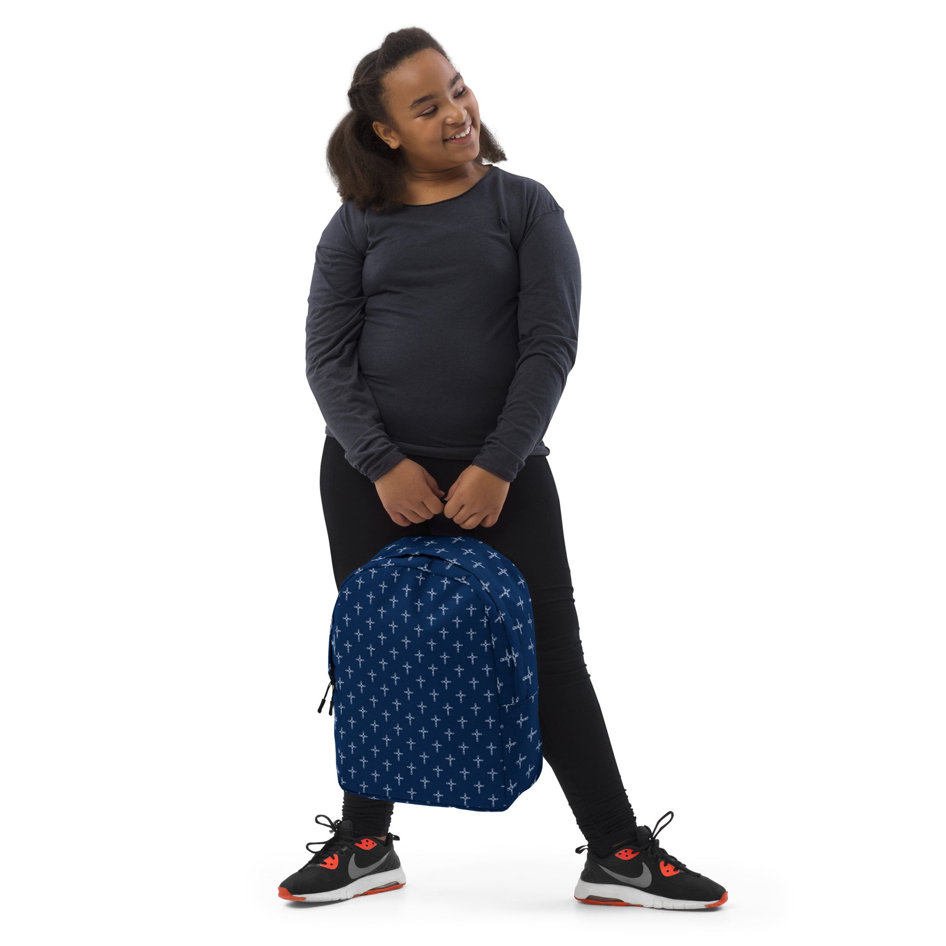 Dark-haired pre-teen girl in dark clothes and tennis shoes holding navy blue backpack that has a white cross print on it.