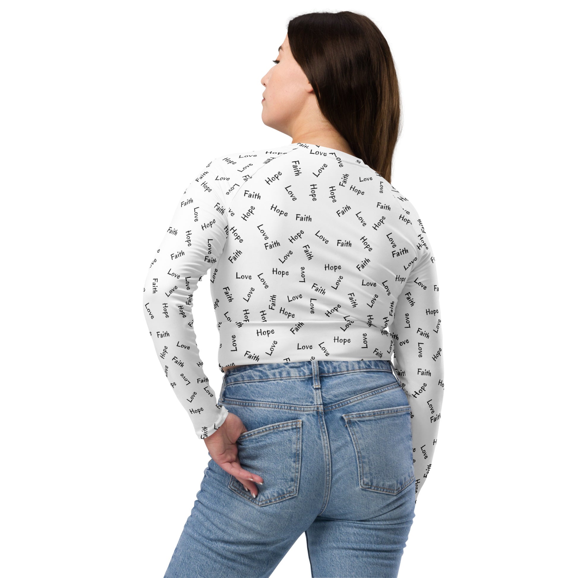 A woman with long dark hair on a white background, wearing light denim blue jeans and a long sleeve white crop top with the words Faith Hope and Love all over it in black print, back view.