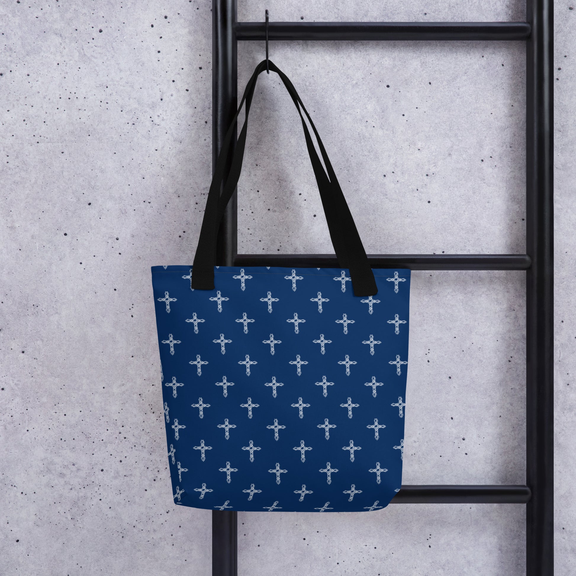 Navy blue tote bag with white crosses on it, hanging on a hook on a black ladder.