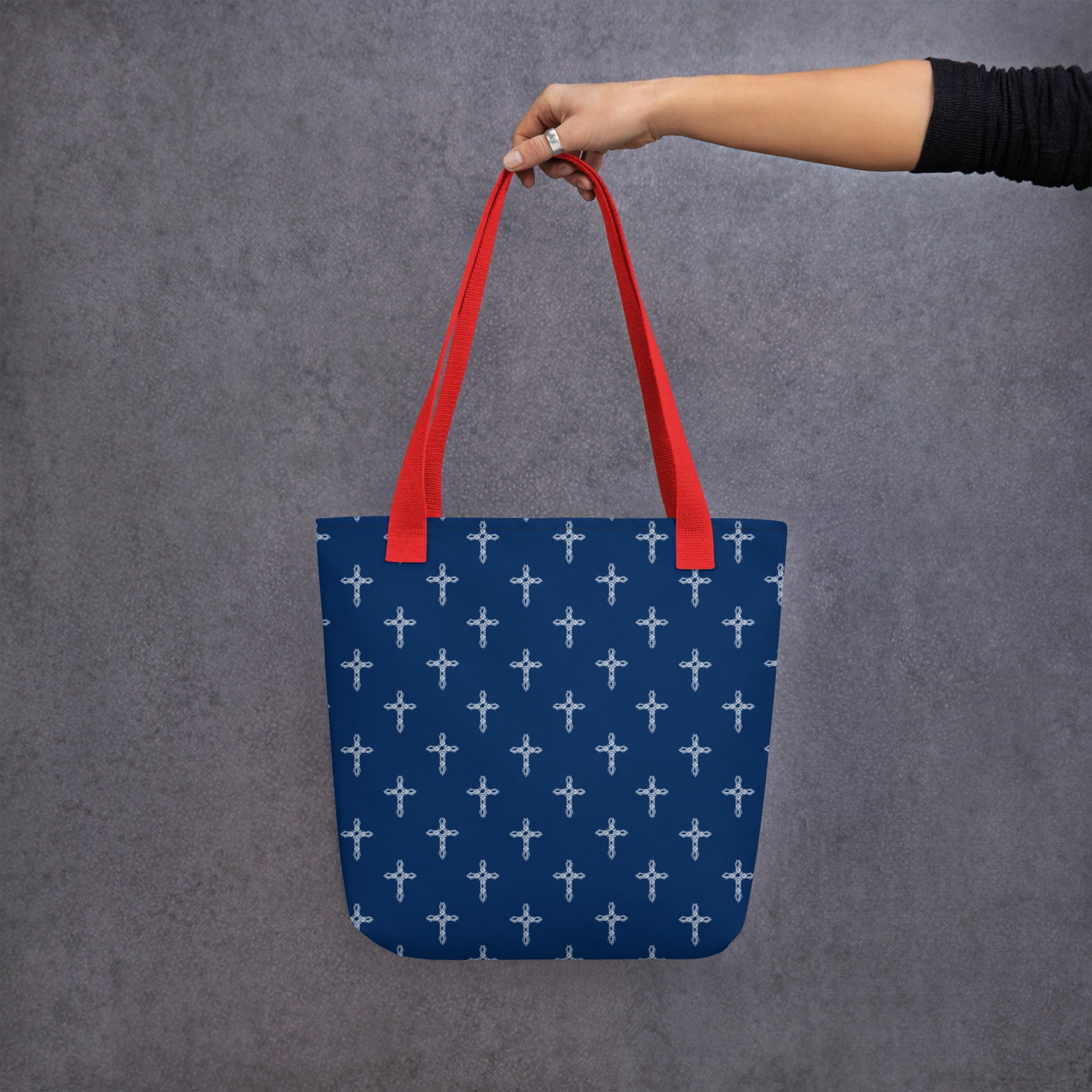 Woman's arm with hand holding the red handle of a navy purse or tote bag with white crosses on it against a neutral gray background.