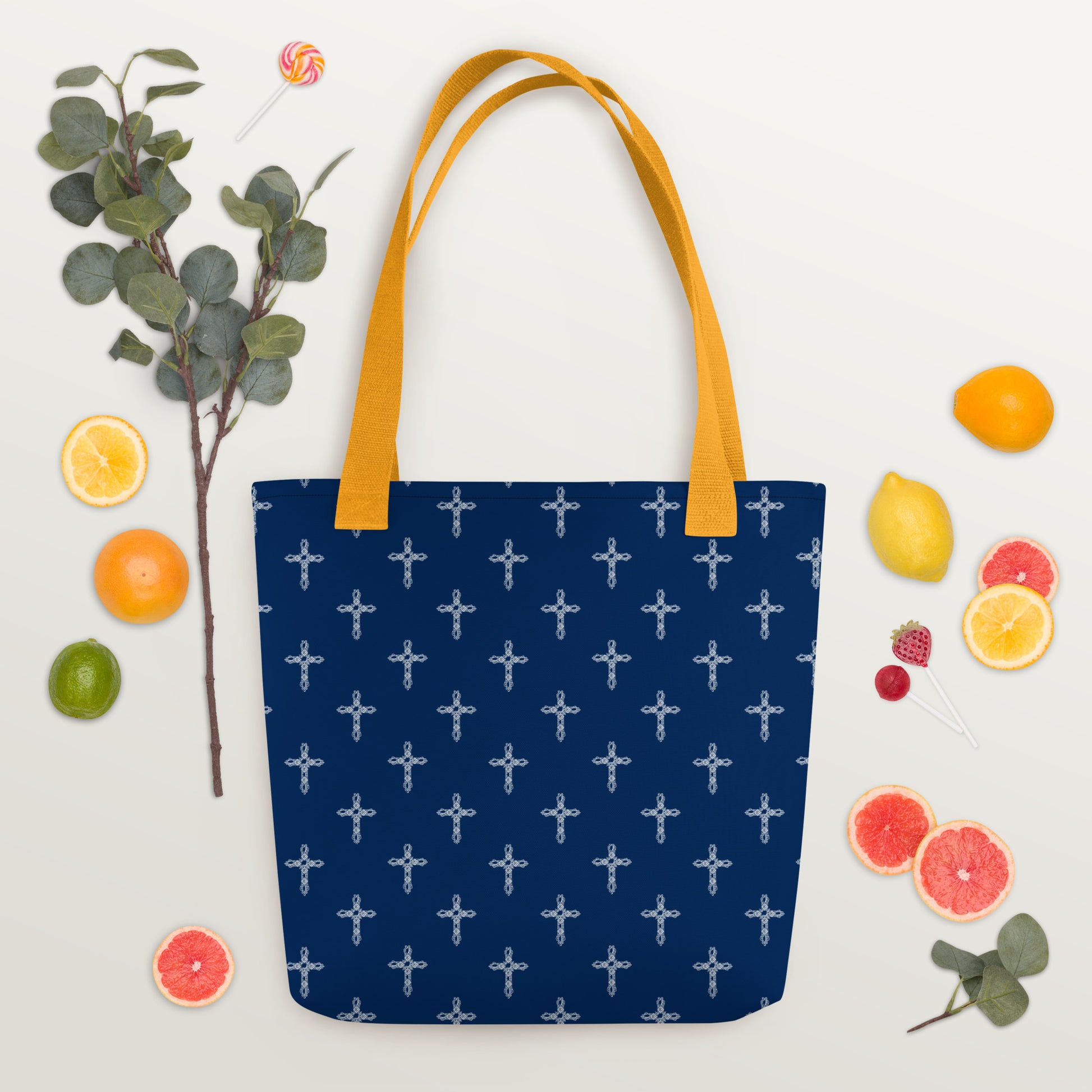 Navy blue purse or bag with white crosses on it and bright yellow handles, laying flat on a table with citrus fruit slices, strawberries, eucalyptus, and a sucker all laying on the white table around the purse.