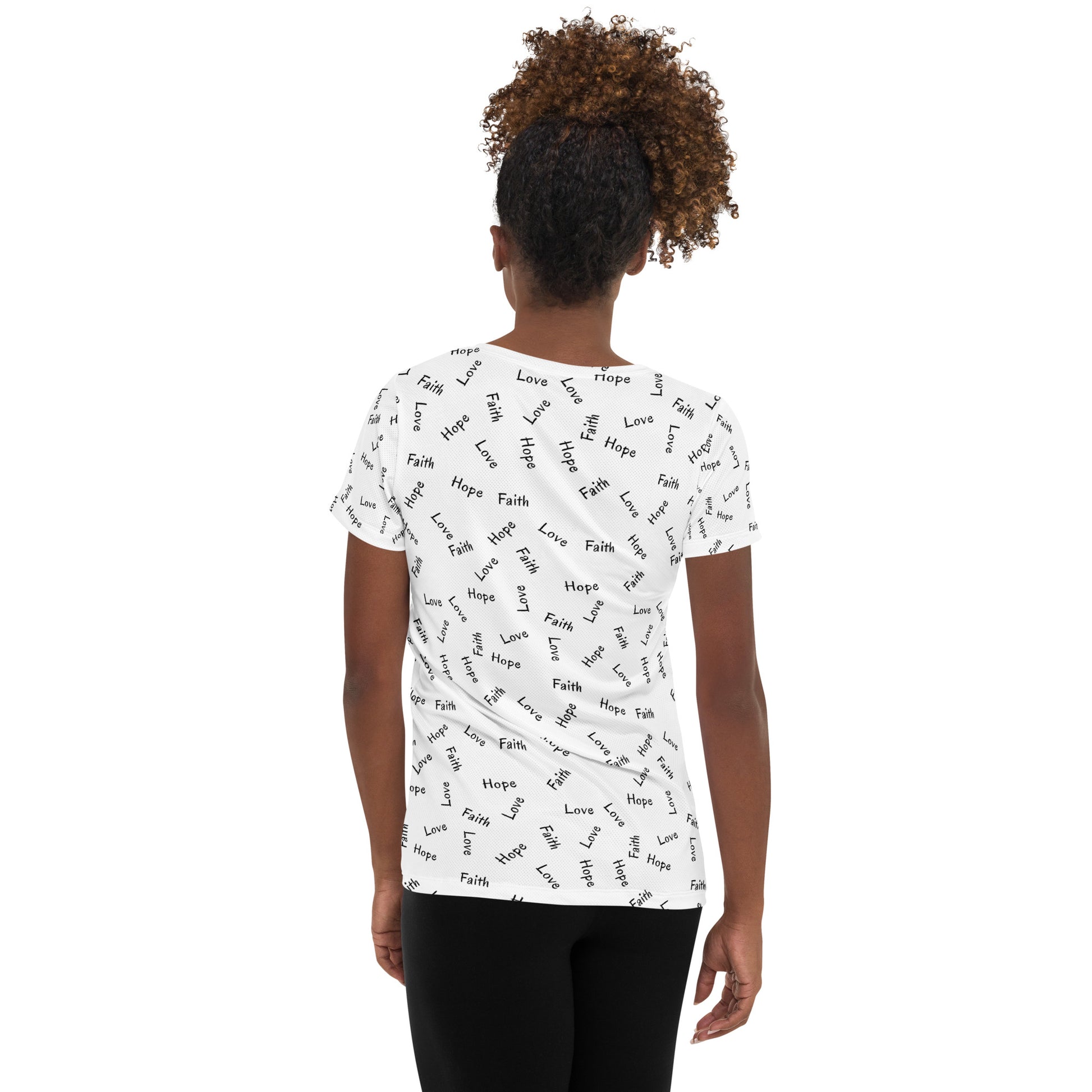 A back view of a woman with long curly hair up in a pony tail wearing black pants and a short-sleeved t-shirt that is white with an all over print of the words Faith Hope and Love.