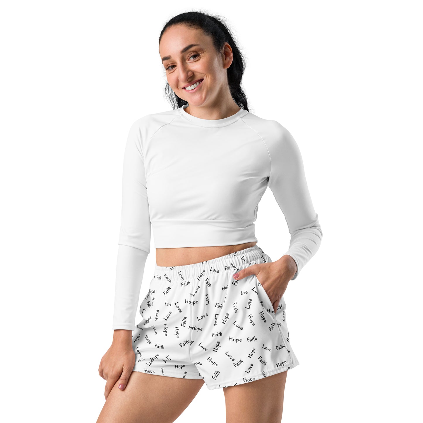 Three-quarter length view of woman with long dark hair wearing a long-sleeve cropped white top and a pair of shorts that are white with an all-over print of Faith Hope and Love words in black, with side pockets and her left hand partially in her left pocket.