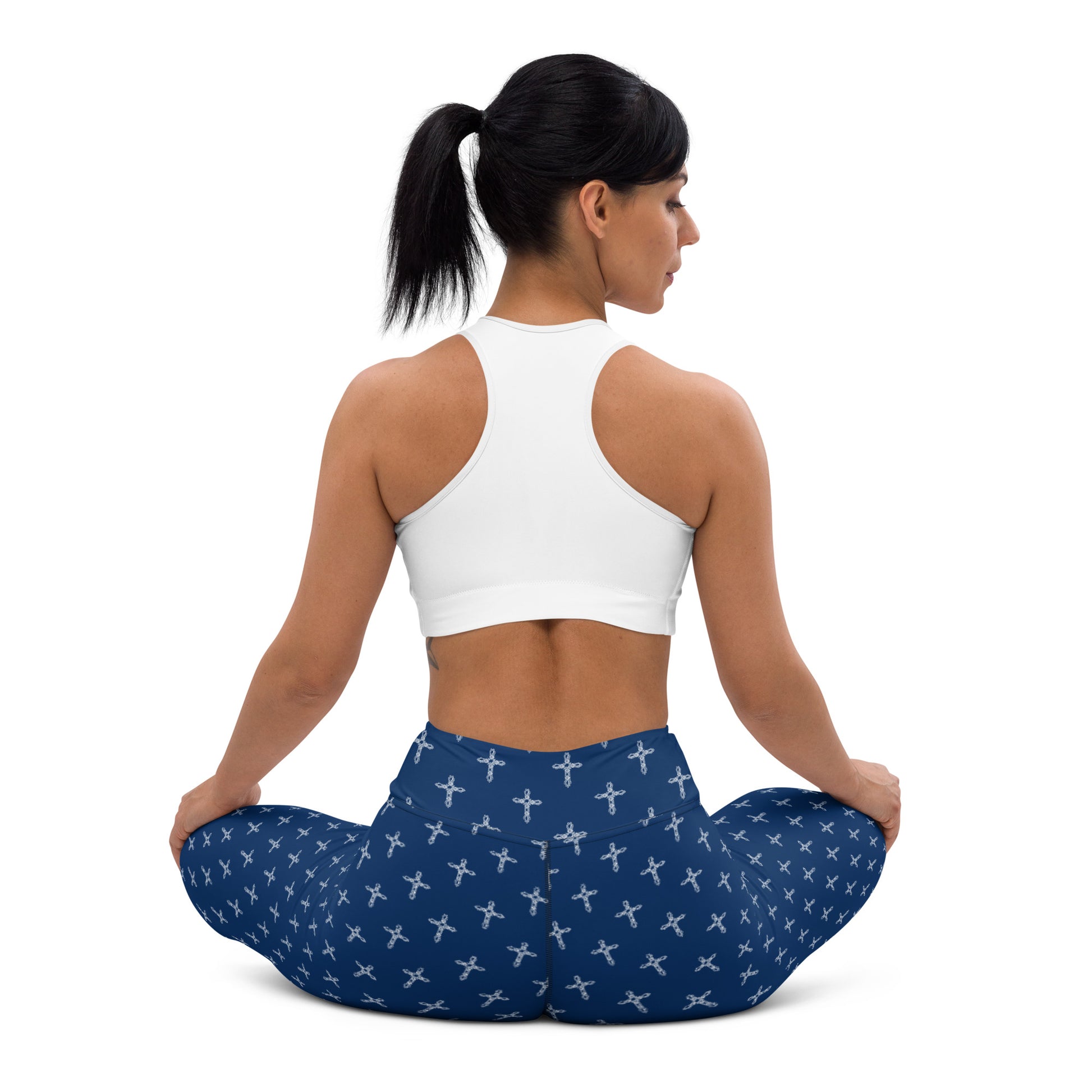 A woman sitting cross-legged on the floor and the view is the woman from the back with her head turned sideways. Her hands are resting on her knees and she has black hair in a ponytail and is wearing a white racer-back sports bra and an pair of high-waisted stretch leggings that are navy with a white cross print all over.