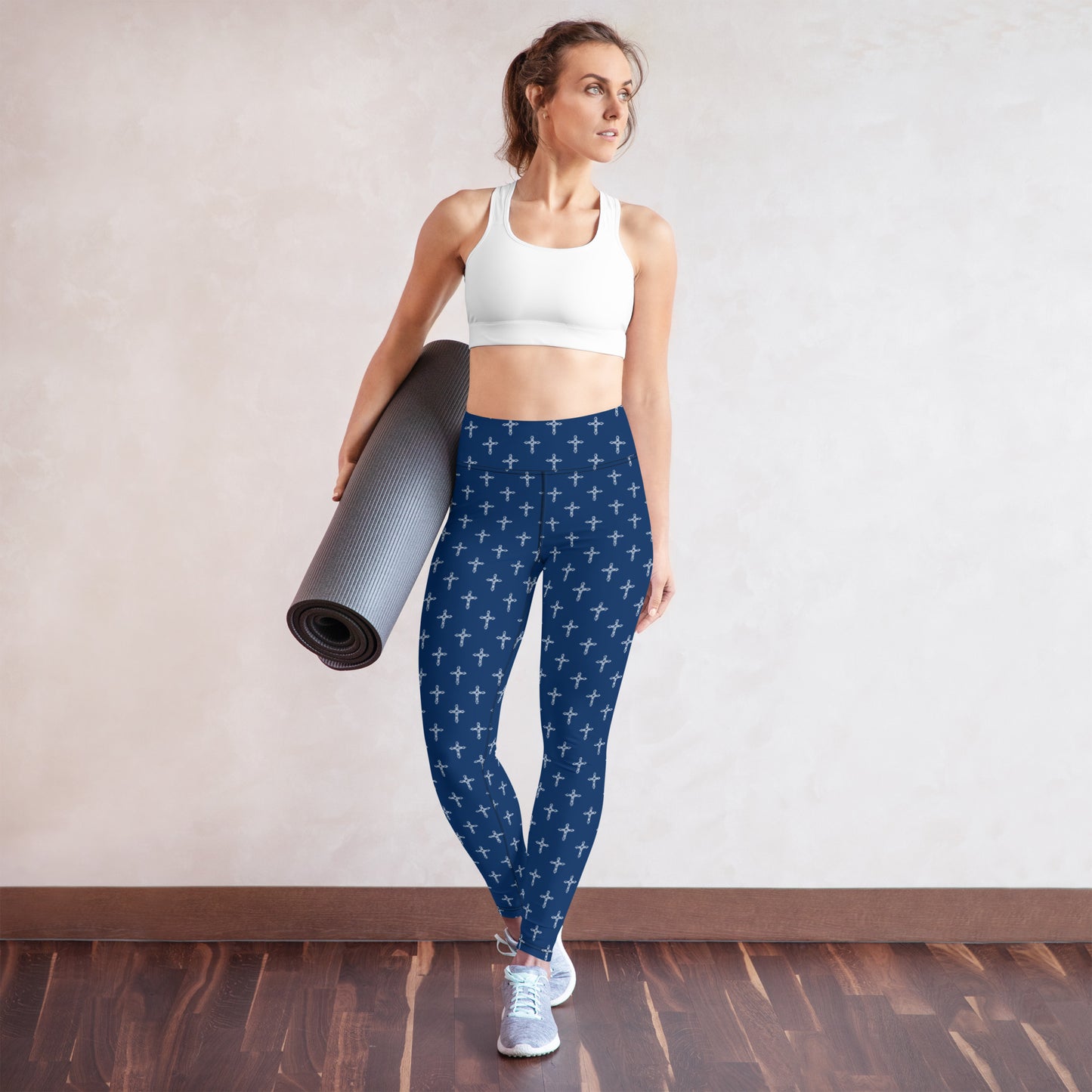 A thin, fit woman with hair in a ponytail holding a gray yoga mat, wearing a white sports bra and gray athletic shoes and a pair of navy leggings with a white cross print all over them, standing on a wooden floor in front of a white wall.