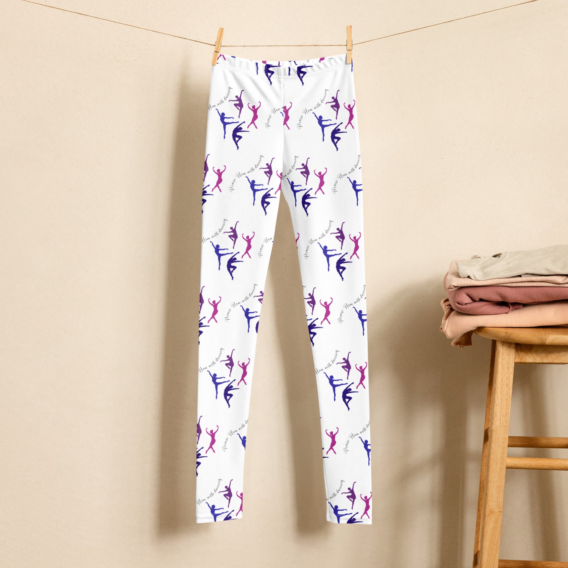 A pair of leggings - white with blue, purple, and pink dancers in different poses and some words - is hanging on a clothesline with clothespins. To the right is a wooden stool with folded clothes on top.