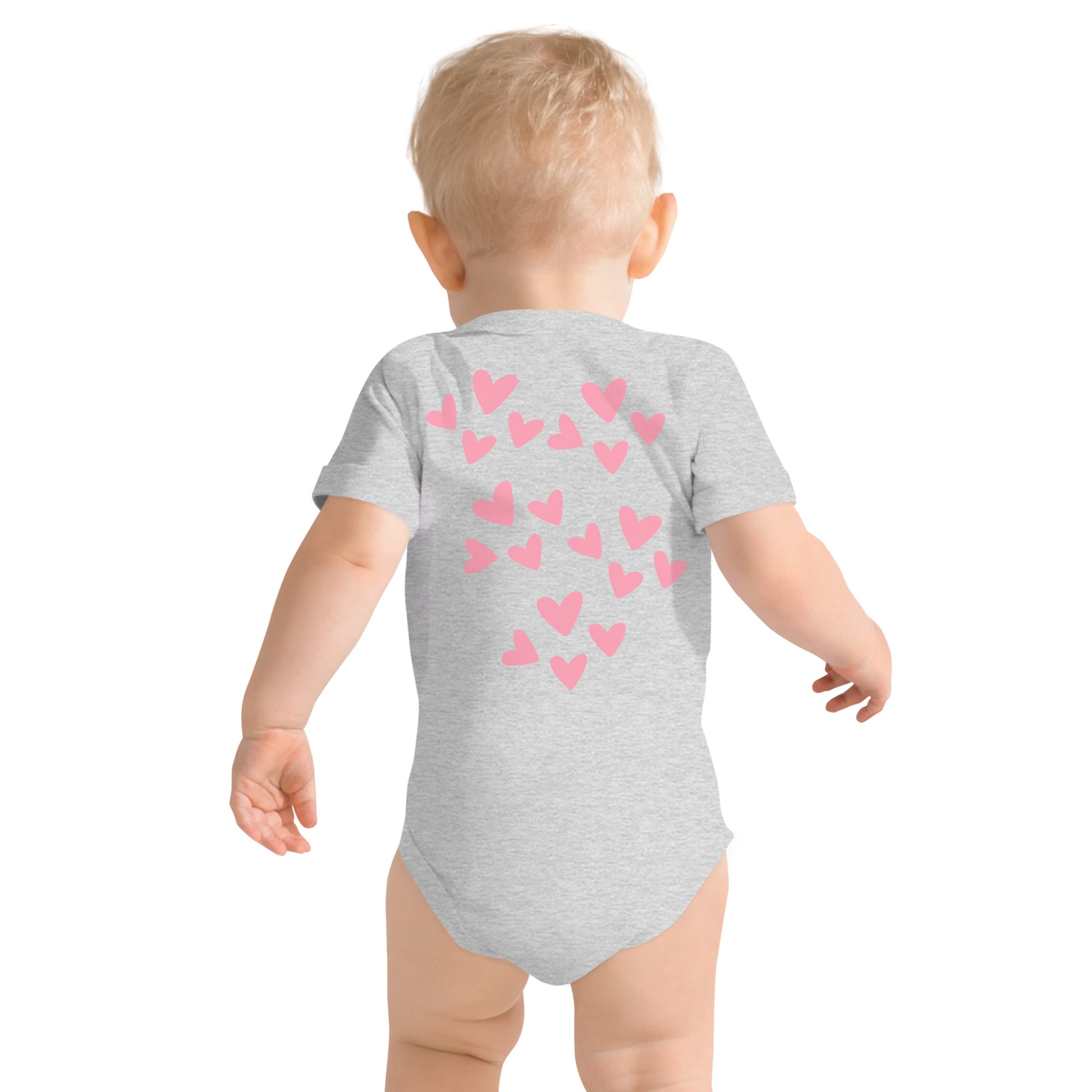 A blonde-haired toddler is walking away and you see the back of the light gray onesie he is wearing, which is covered in pink hearts.