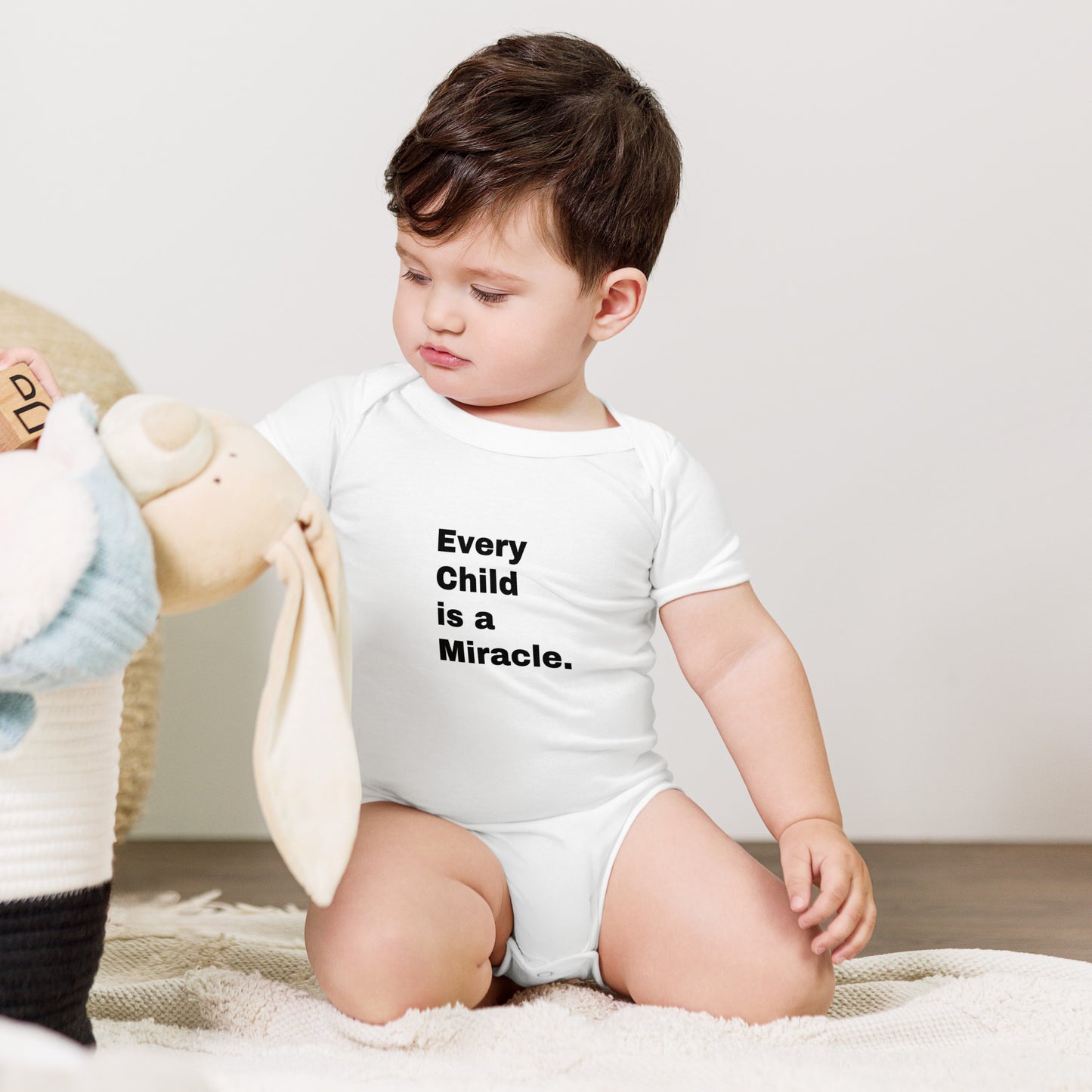 Little boy with dark hair playing on the floor wears a white short-sleeved onesie that says Every Child is a Miracle on the front in black text.