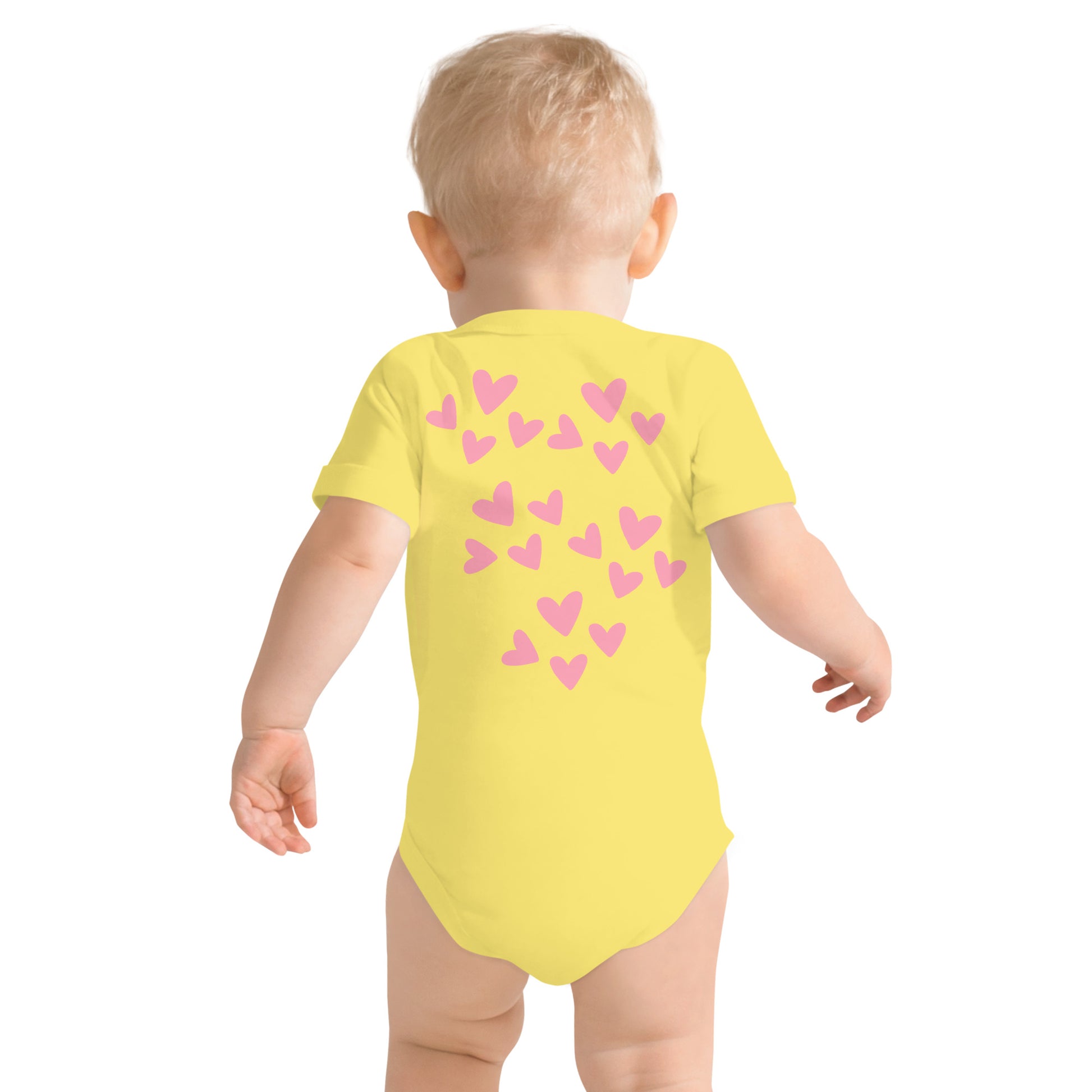 A blonde toddler is facing away from the camera and we see his back and the back of the yellow short-sleeved onesie he is wearing, which is covered with light pink hearts of varying sizes.