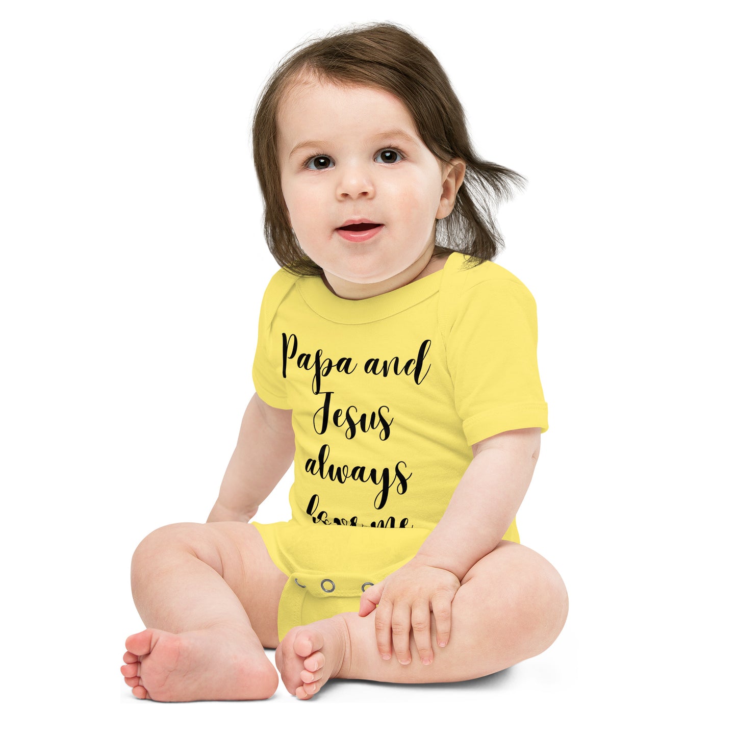 A toddler is seated on a white background wearing a yellow short-sleeved onesie that says Papa and Jesus always love me in script.