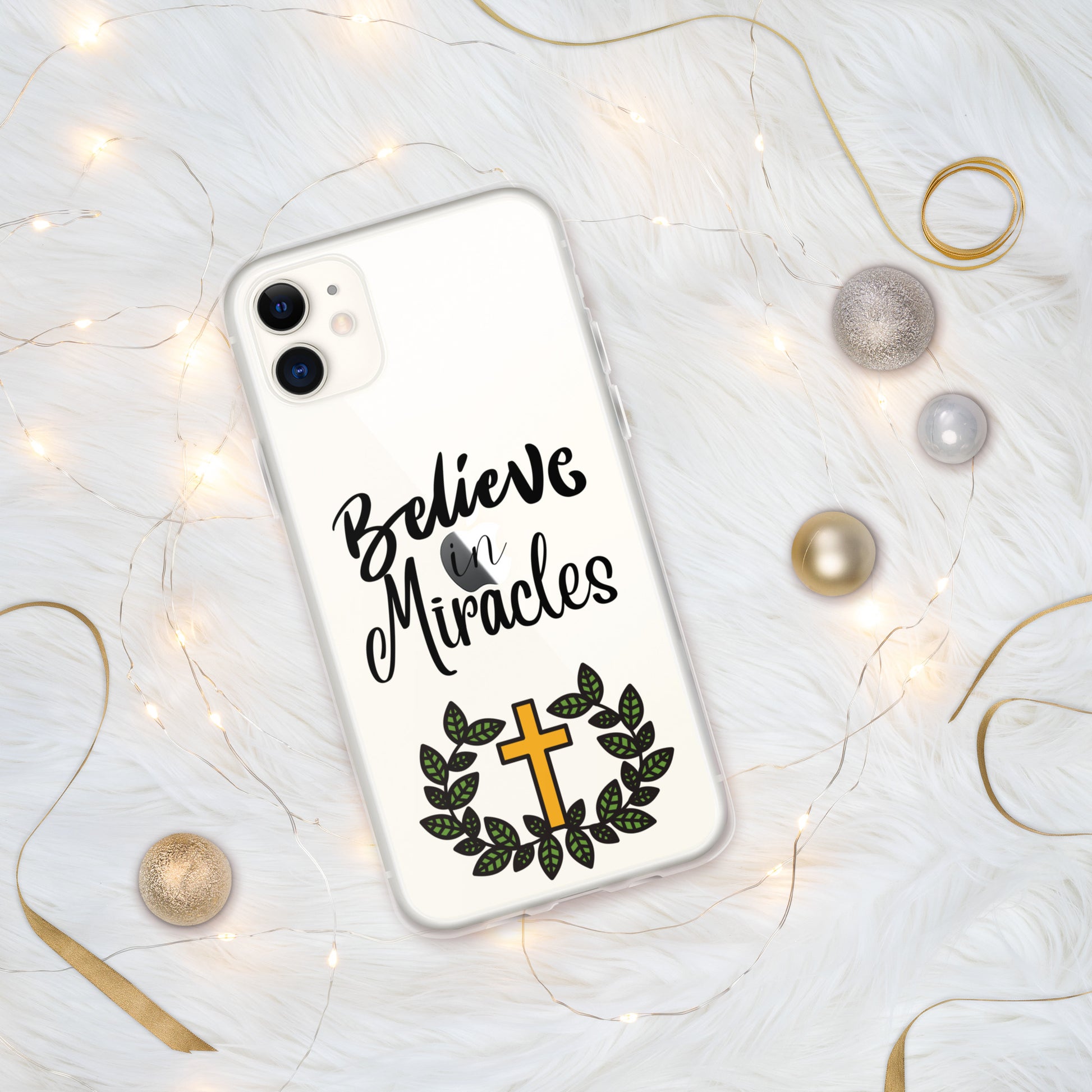 Clear iphone case on white iphone surrounded by little lights and gold ribbons and round gold and silver balls. The phone case has a gold cross and green leaf wreath at the bottom and Believe in Miracles word art in the middle.