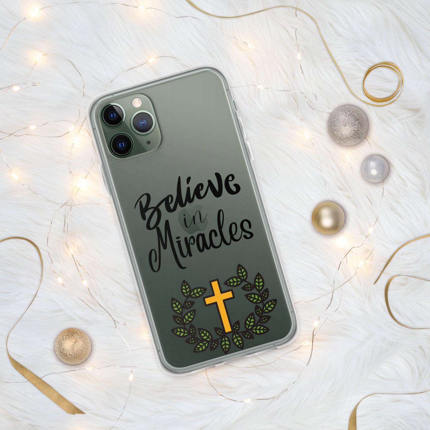 Clear iphone cover on a dark gray or green iphone with the words "Believe in Miracles" in fancy lettering in the middle and at the bottom a gold cross and green leaves encircled around it and it is on a white table with mini lights and gold ribbons and gold and silver balls around it.