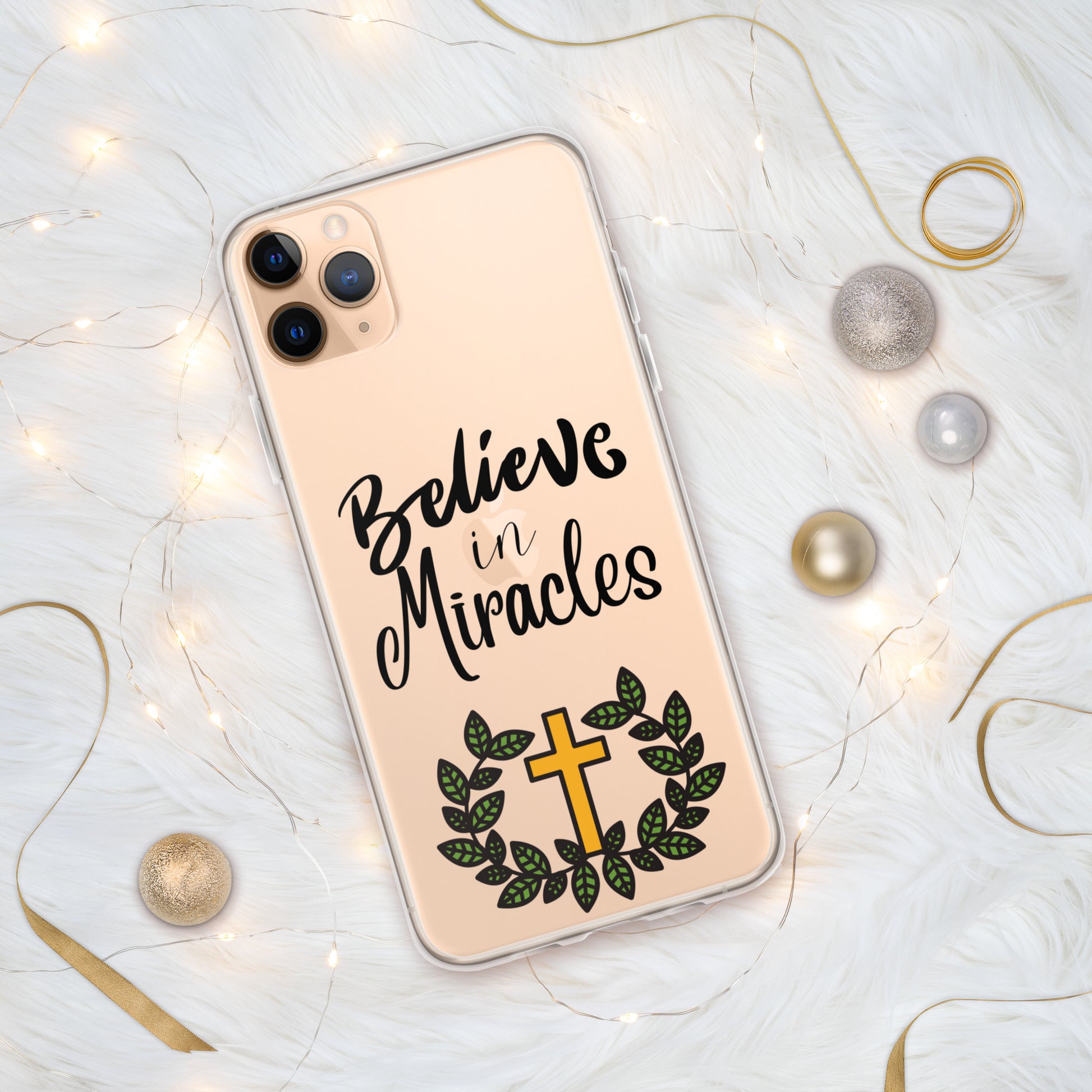 Clear phone case on a gold iphone that says Believe in Miracles in ornate text over a gold cross with green leaves around it like a wreath and ornate lights and ribbons and baubles around the phone.