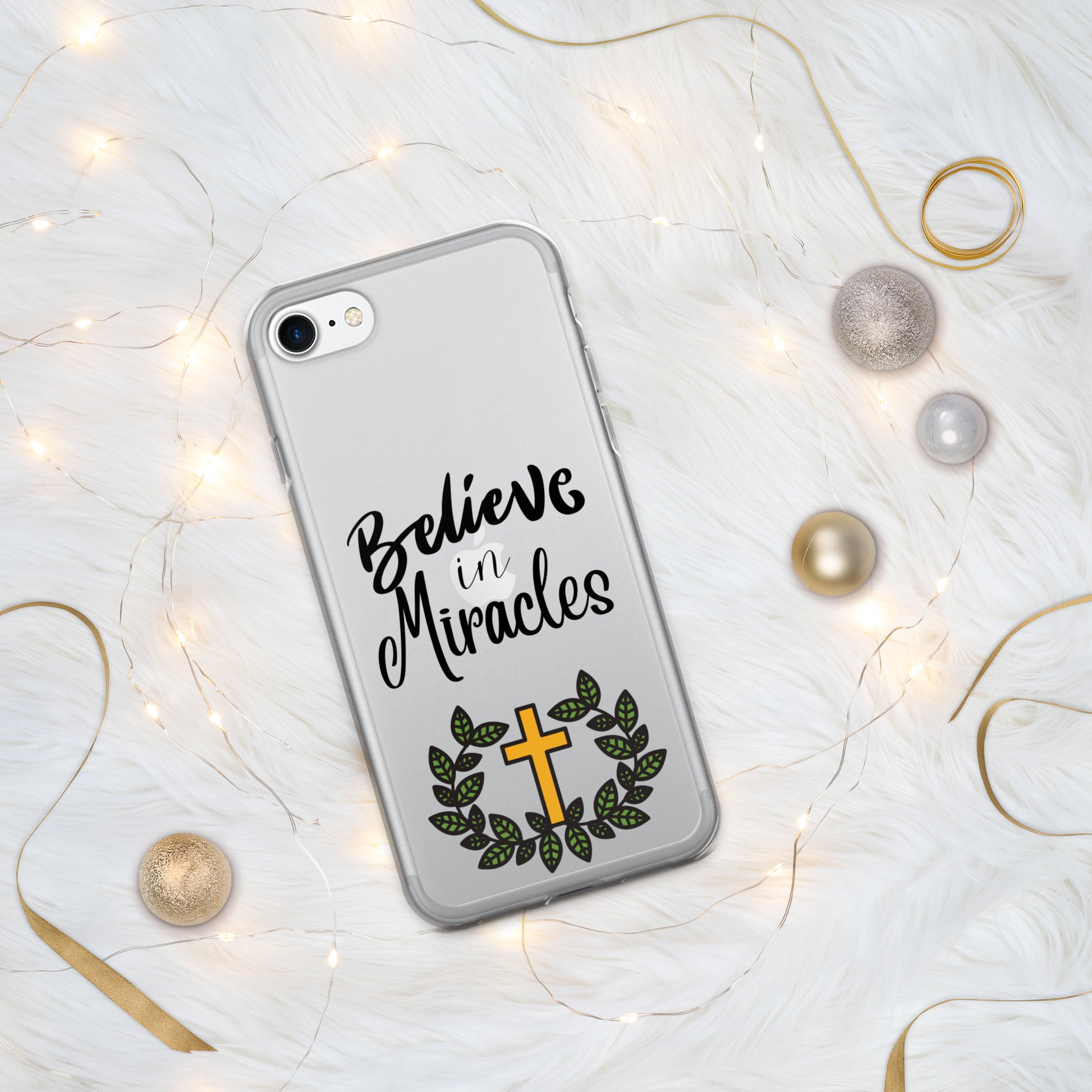 Clear iphone case on a silver cell phone says Believe in Miracles in ornate letters and has a gold cross and green leaf wreath beneath it, with tiny white lights, gold curled ribbons, and gold and silver balls around it.