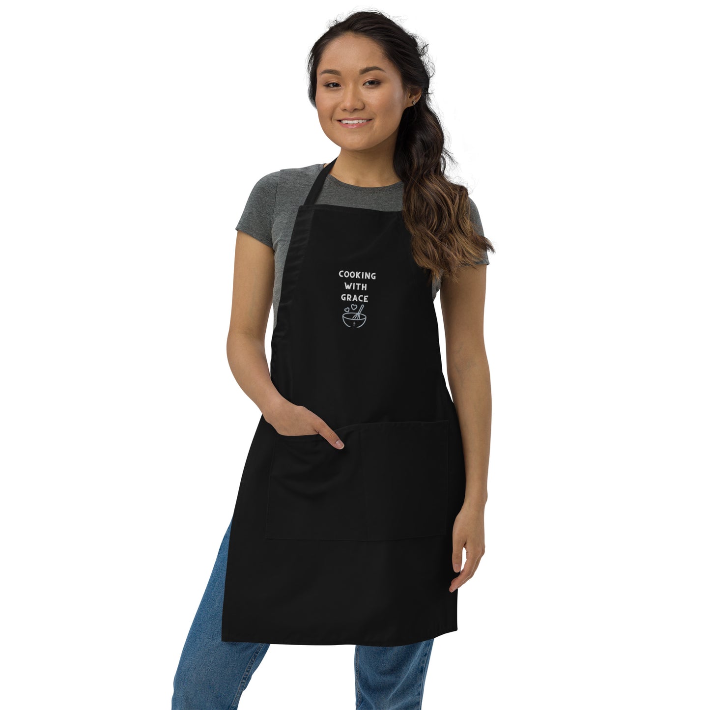 Dark haired smiling woman wearing jeans and short-sleeve gray t-shirt, over which she is wearing a black apron, shown full length, that is embroidered with COOKING WITH GRACE, two hearts, a bowl, and a whisk, and the woman has one hand in the front pocket of the apron.