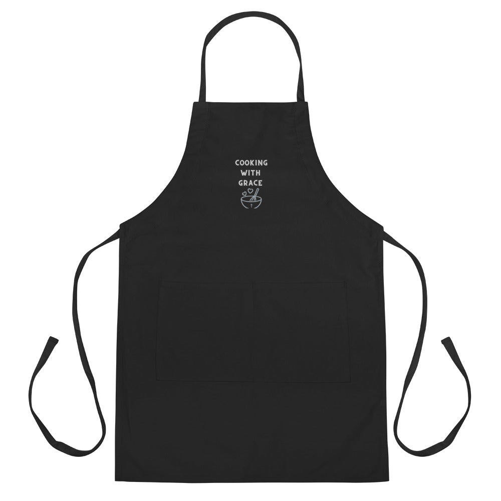 Black apron on plain white background showing full length, ties, and embroidered front with graphic and the words:  COOKING WITH GRACE.