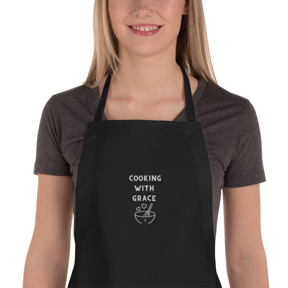 Middle section of smiling blonde woman wearing brown short-sleeve t-shirt and black apron embroidered with Cooking with Grace and bowl, whisk, and two hearts.