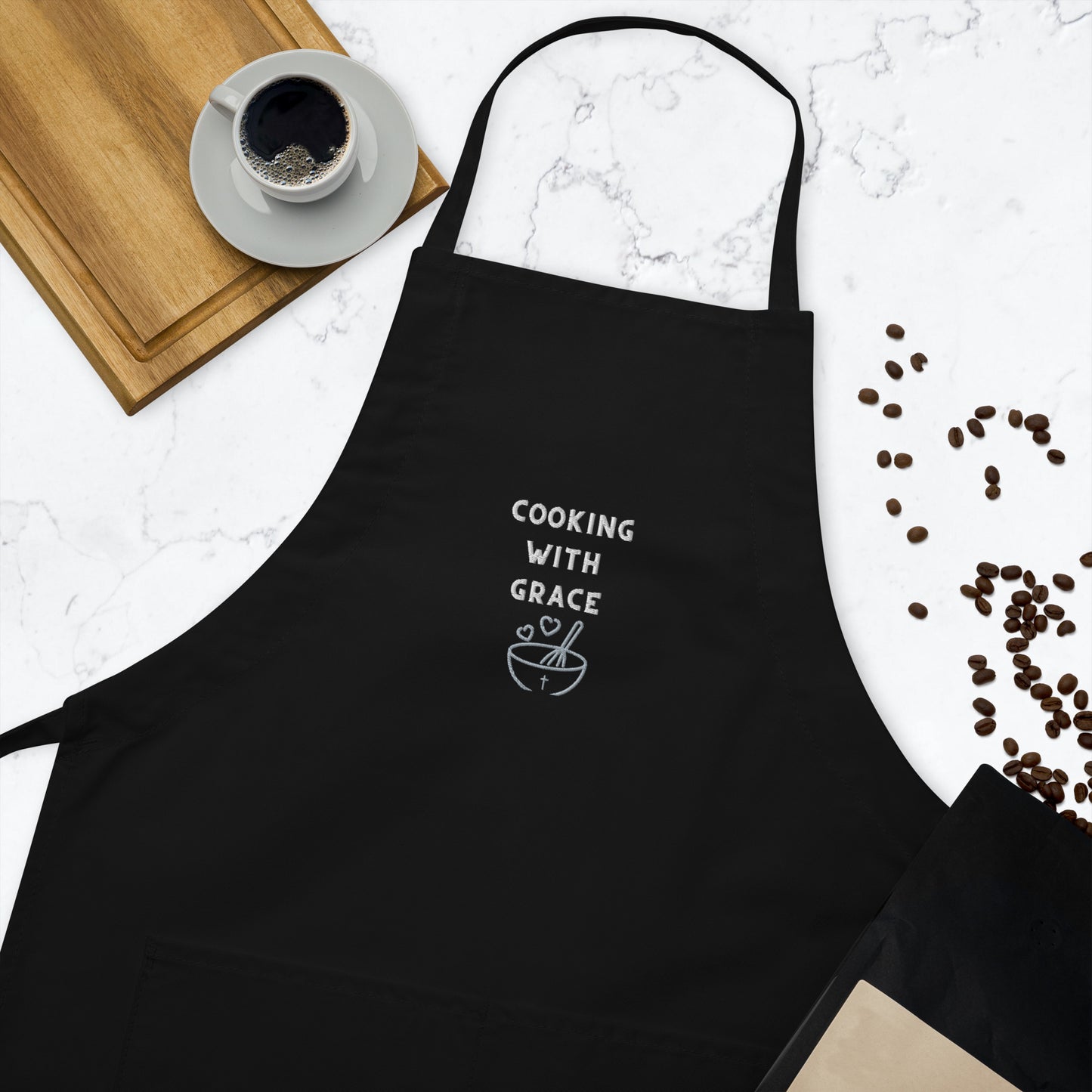 Cuting board, cup of coffee on white saucer, and coffee beans scattered around black apron embroidered with Cooking With Grace and a mixing bowl, whisk, and two hearts.