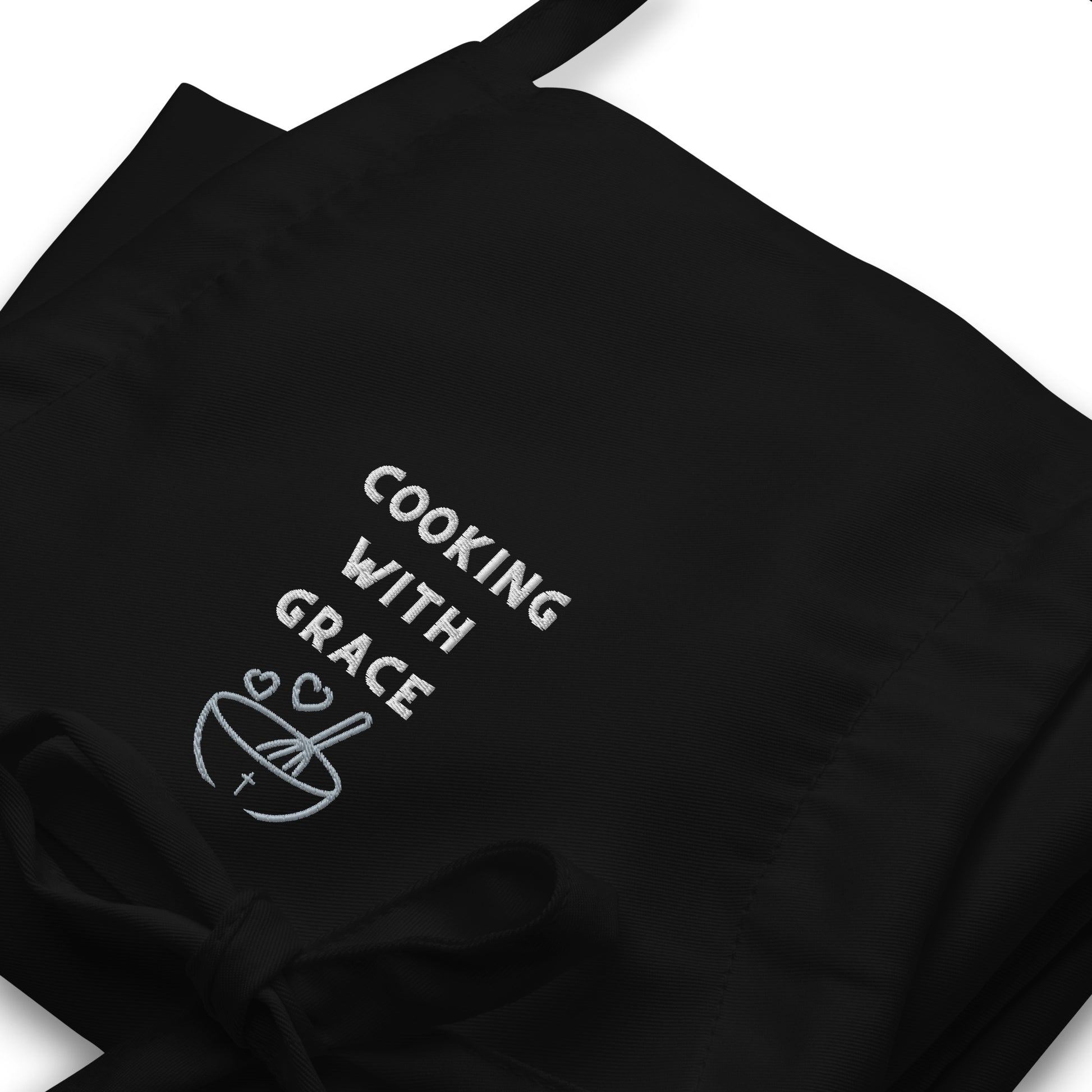 Folded black apron showing close up of embroidered front that says COOKING WITH GRACE and a mixing bowl with a cross on it, two hears, and a whisk in the bowl.