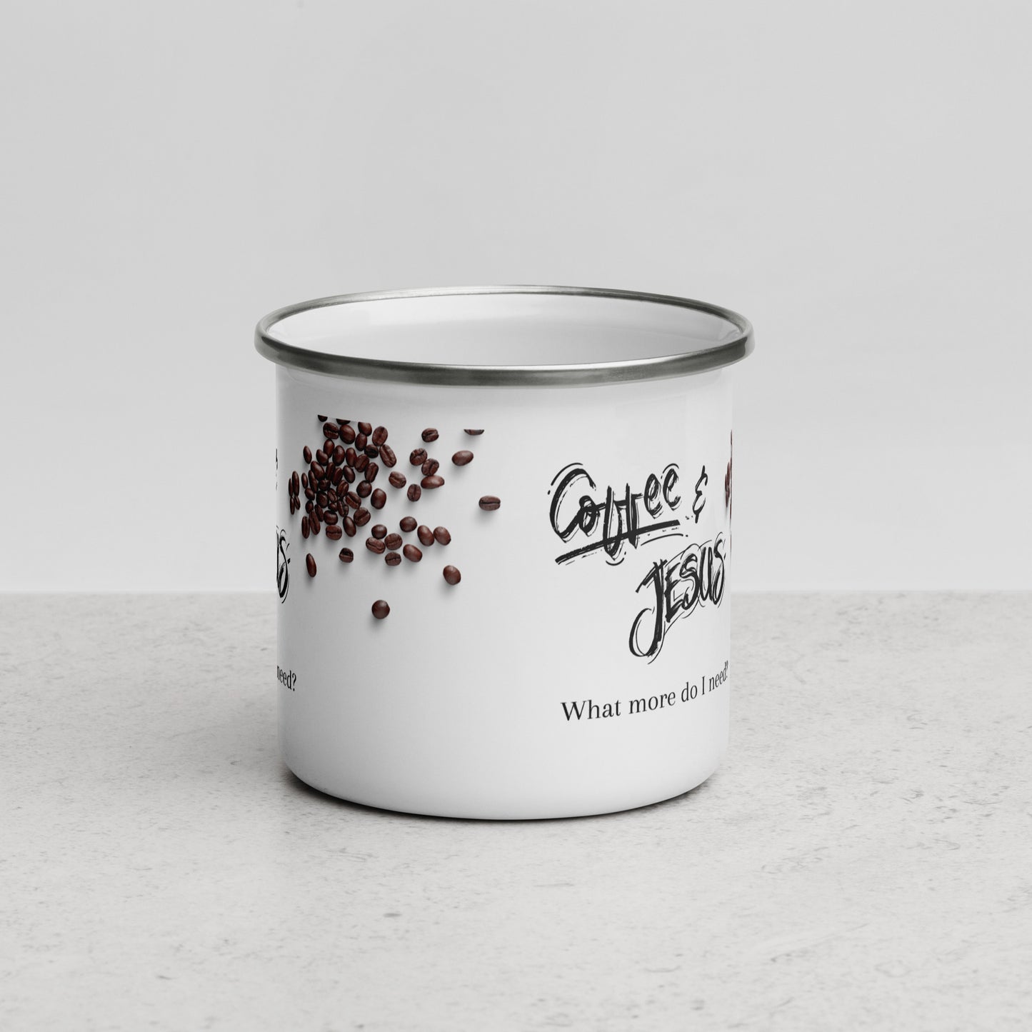 White enamel metal coffee mug has a picture of scattered coffee beans and the words Coffee & Jesus and "What more do I need" on the side in two different black fonts, sitting on a whitish counter top with a white background.