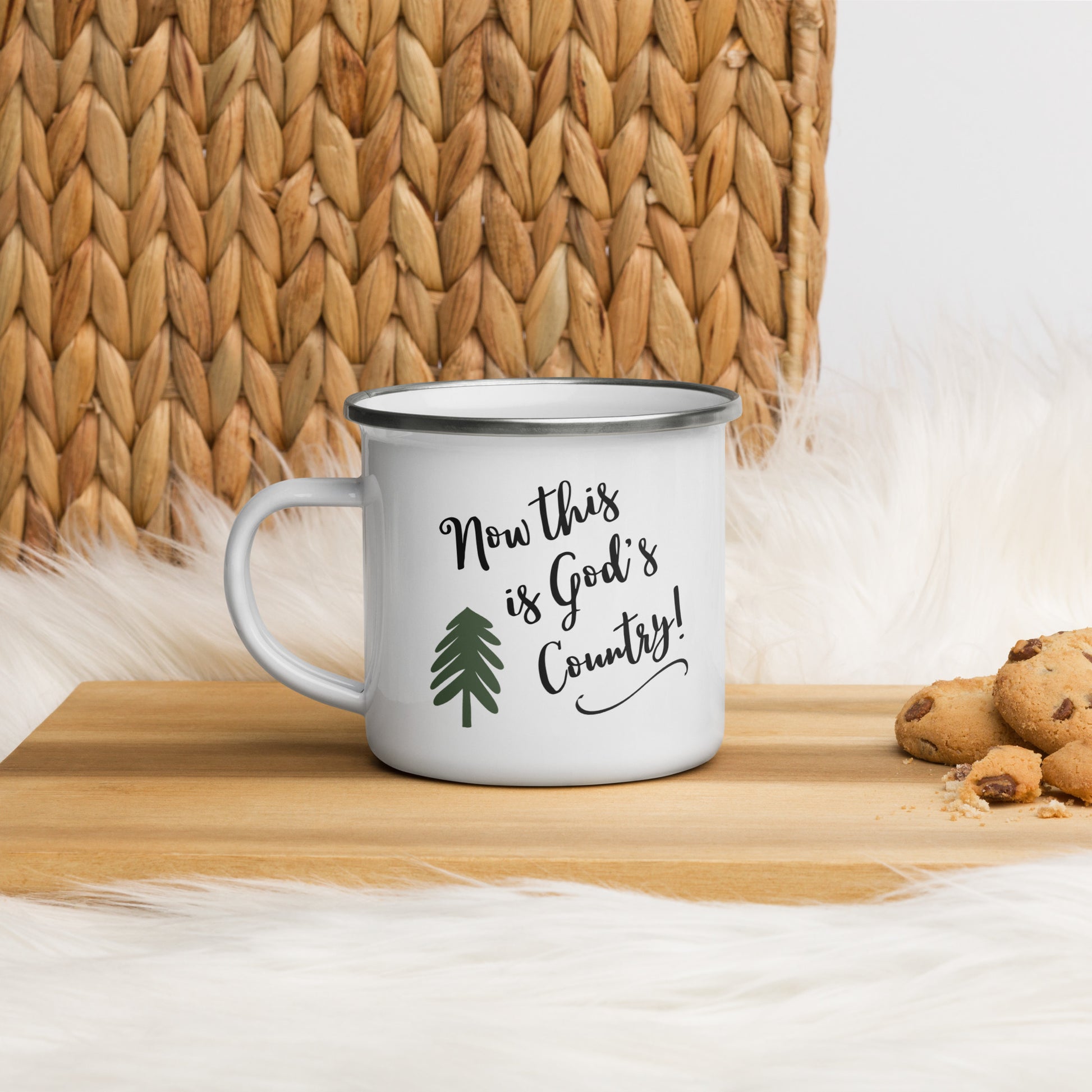 A wooden cutting board sits on a white fluffy fur with a woven tan textile in the background. On the cutting board, there is a white enamel mug that has a green pine tree on it and the words:  Now this is God's country! in script with a swoosh under it and some chocolate chip cookies on it to the far right.