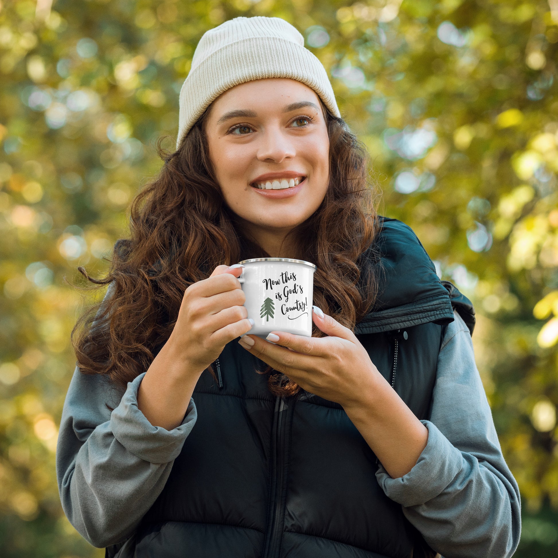 A smiling woman with long dark curly hair is outdoors, dressed in a blue shirt with rolled up sleeves, and a navy sleeveless vest, and she holds a white enamel coffee mug with a green pine tree on it and the words:  Now this is God's Country!