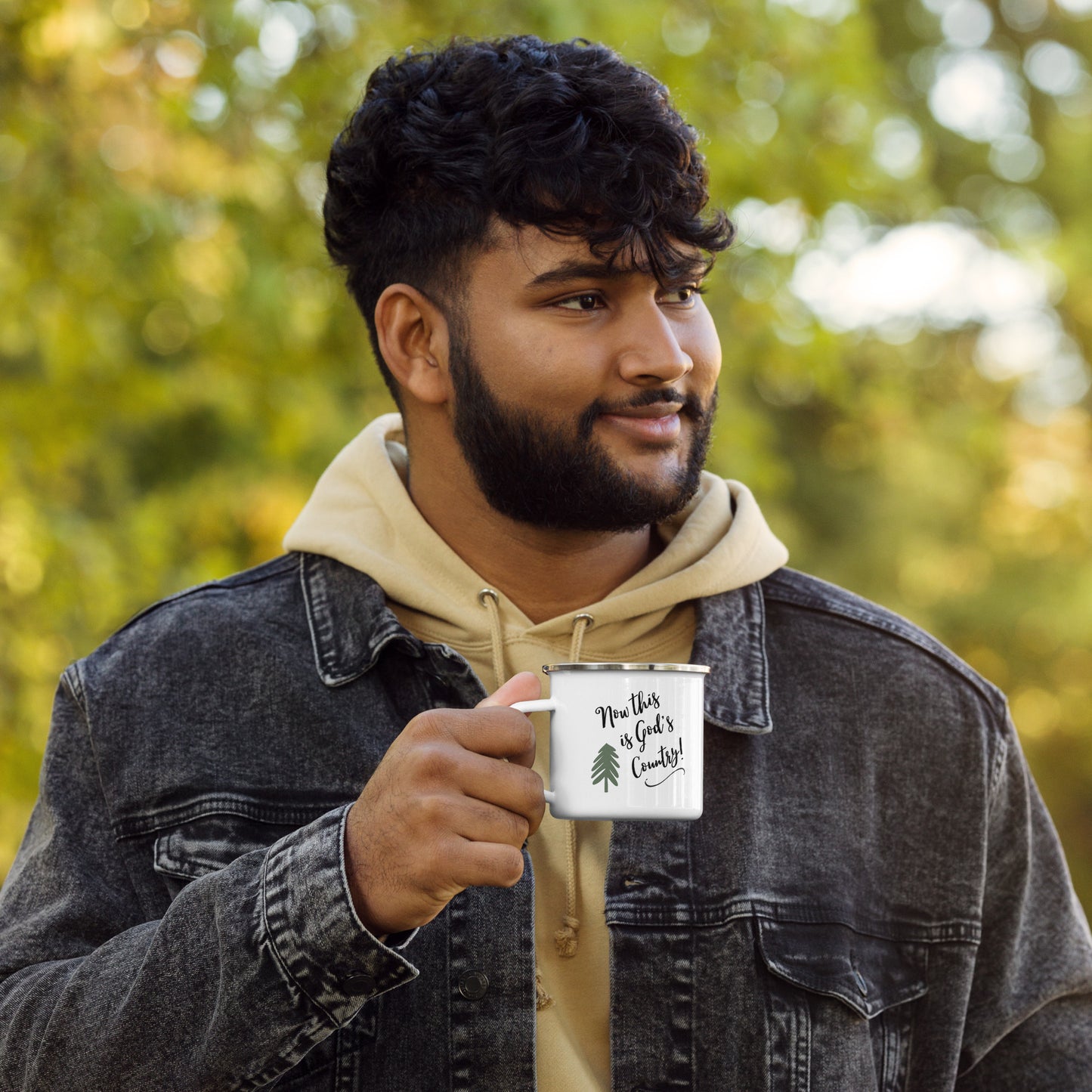 A dark-haired man with a dark beard and mustache is outdoors looking off in the distance. He wears a tan hooded sweatshirt and a black denim jacket over top and holds a white enamel mug that says Now this is God's Country and has a simple green  pine tree on it.