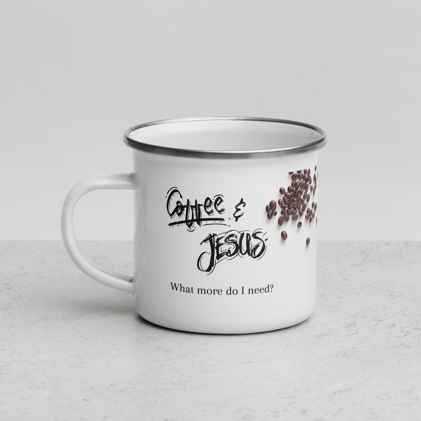 White enamel or metal coffee mug with a silver edge has the words "Coffee & Jesus What more do I need?" printed on the side in two different fonts, with coffee beans on the side and it is sitting on a white countertop with a white background.