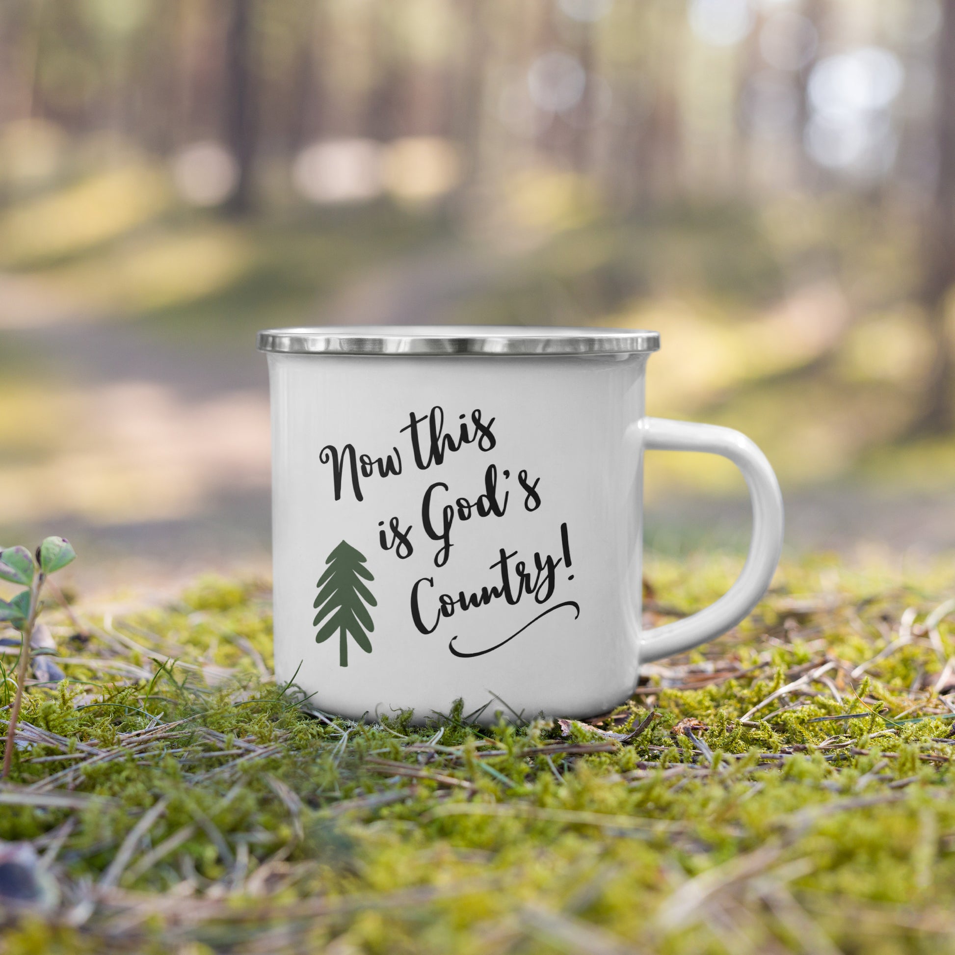 Outdoors, on a section of green moss covered ground with twigs and pine needles, and a dirt road and trees blurred in the background, sits a single white enamel coffee mug. On it, in script writing, is printed:  Now this is God's Country! It also has a swirl below the letters and a green hand drawn pine tree on the mug.