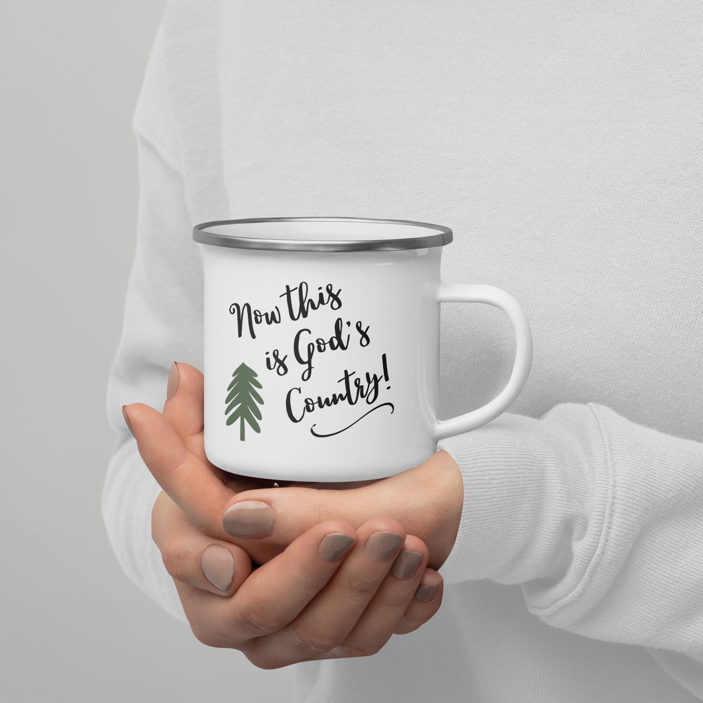 Woman's manicured hands shown holding a white enamel mug and she is wearing a plain white sweatshirt, but you only see the front of the person and her hands. The mug has a simple green pine tree on it and the words:  Now this is God's Country!