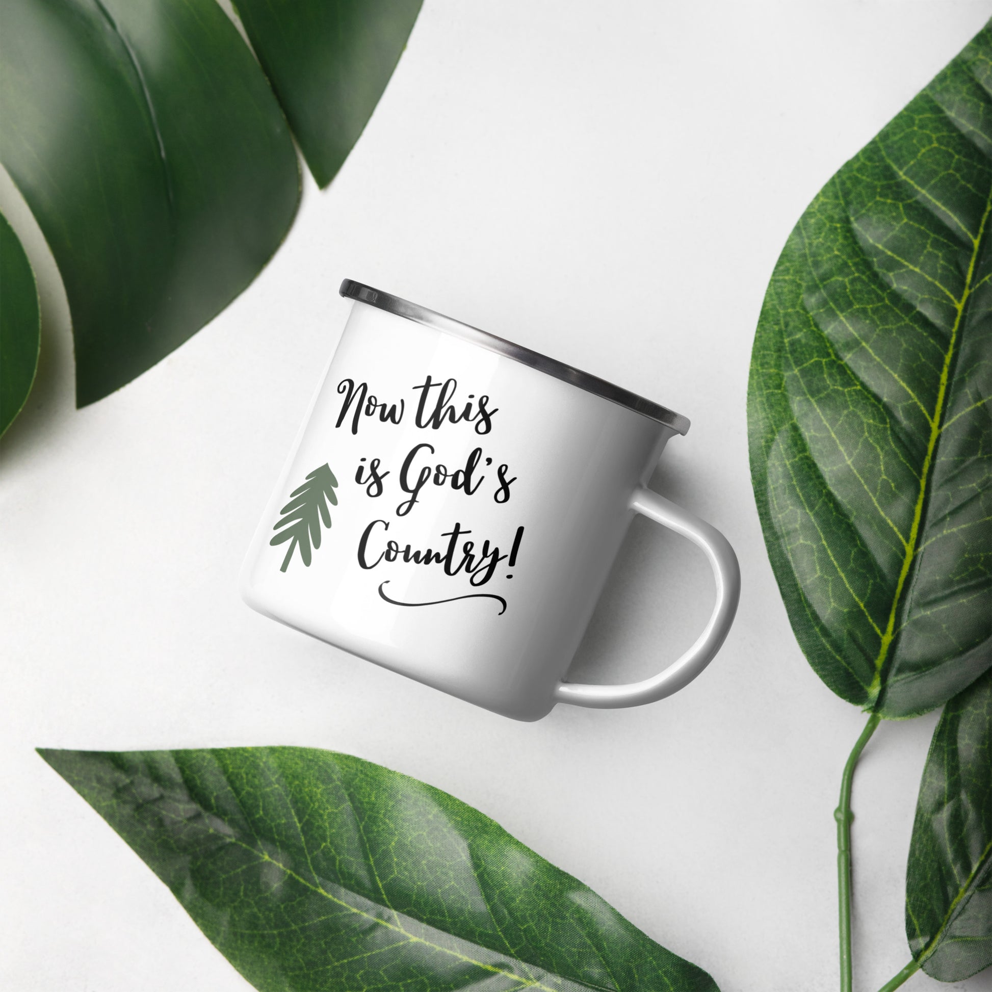 White enamel coffee mug in the center says Now this is God's country and has a hand-drawn pine tree on it. Large green leaves are around the mug on a white background. 