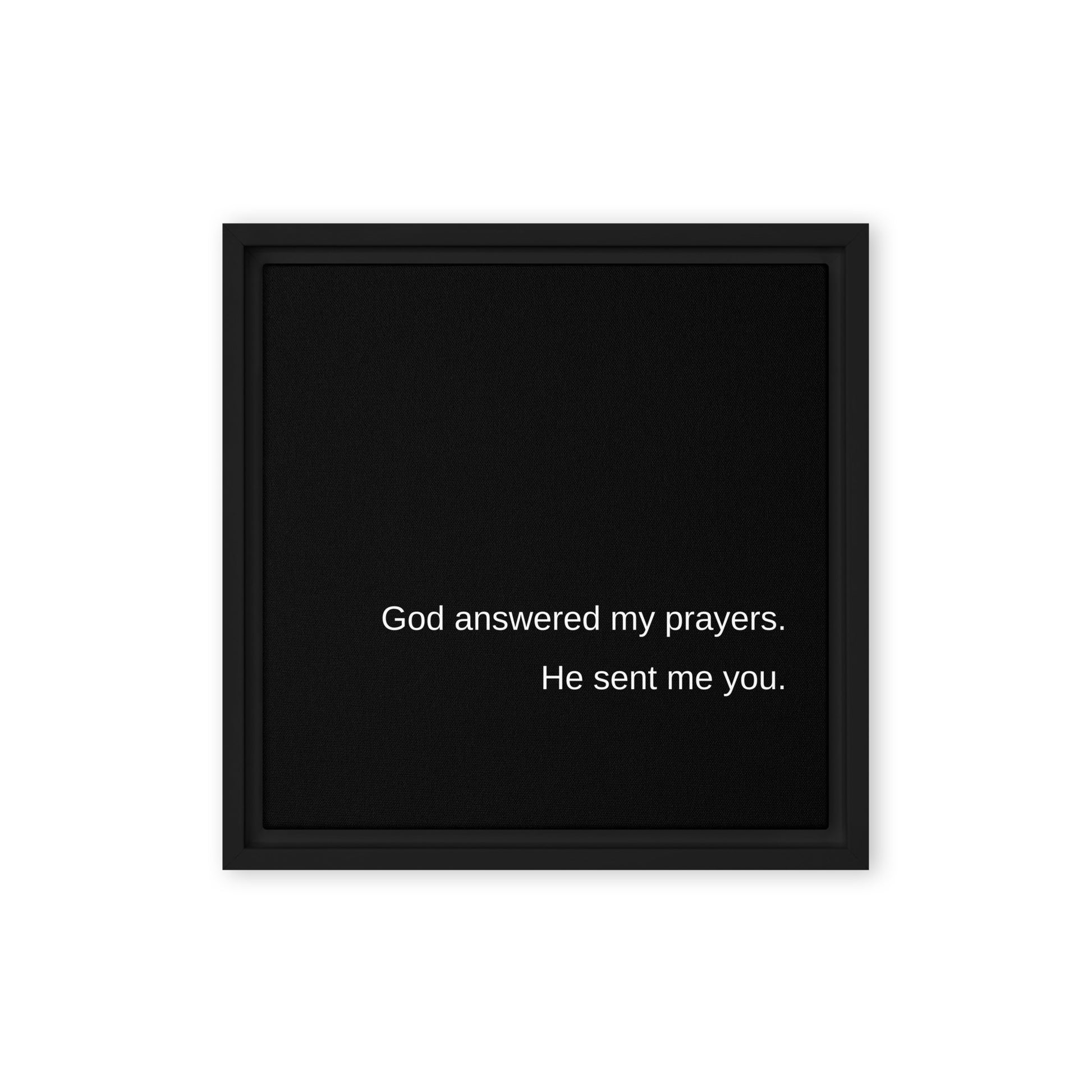 A solid white wall with a square black stretched canvas with a black frame and white text on the black canvas saying: God answered my prayers. He sent me you.