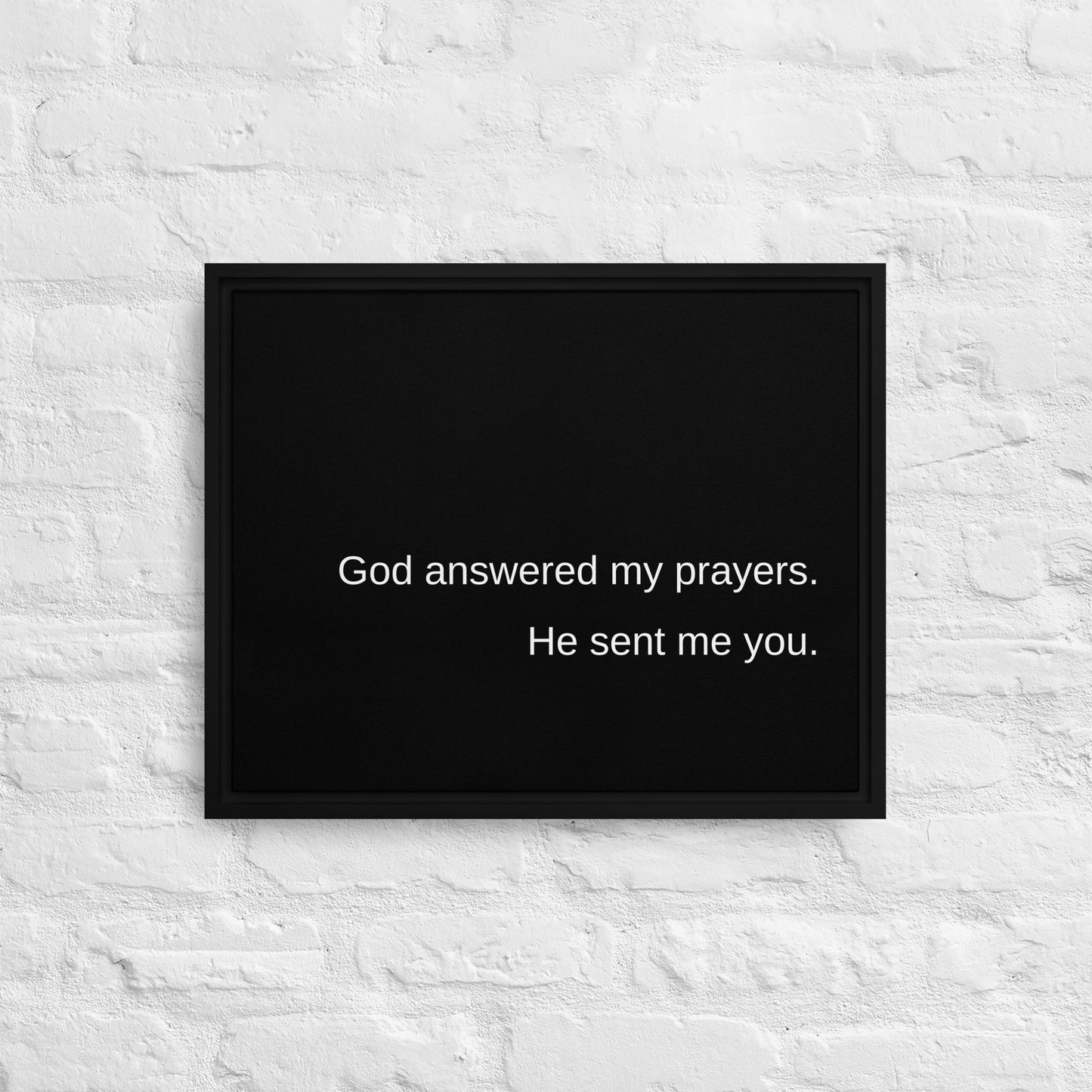 White brick wall with black stretched canvas also framed in black and on the front in white text it says simply:  God answered my prayers. He sent me you.