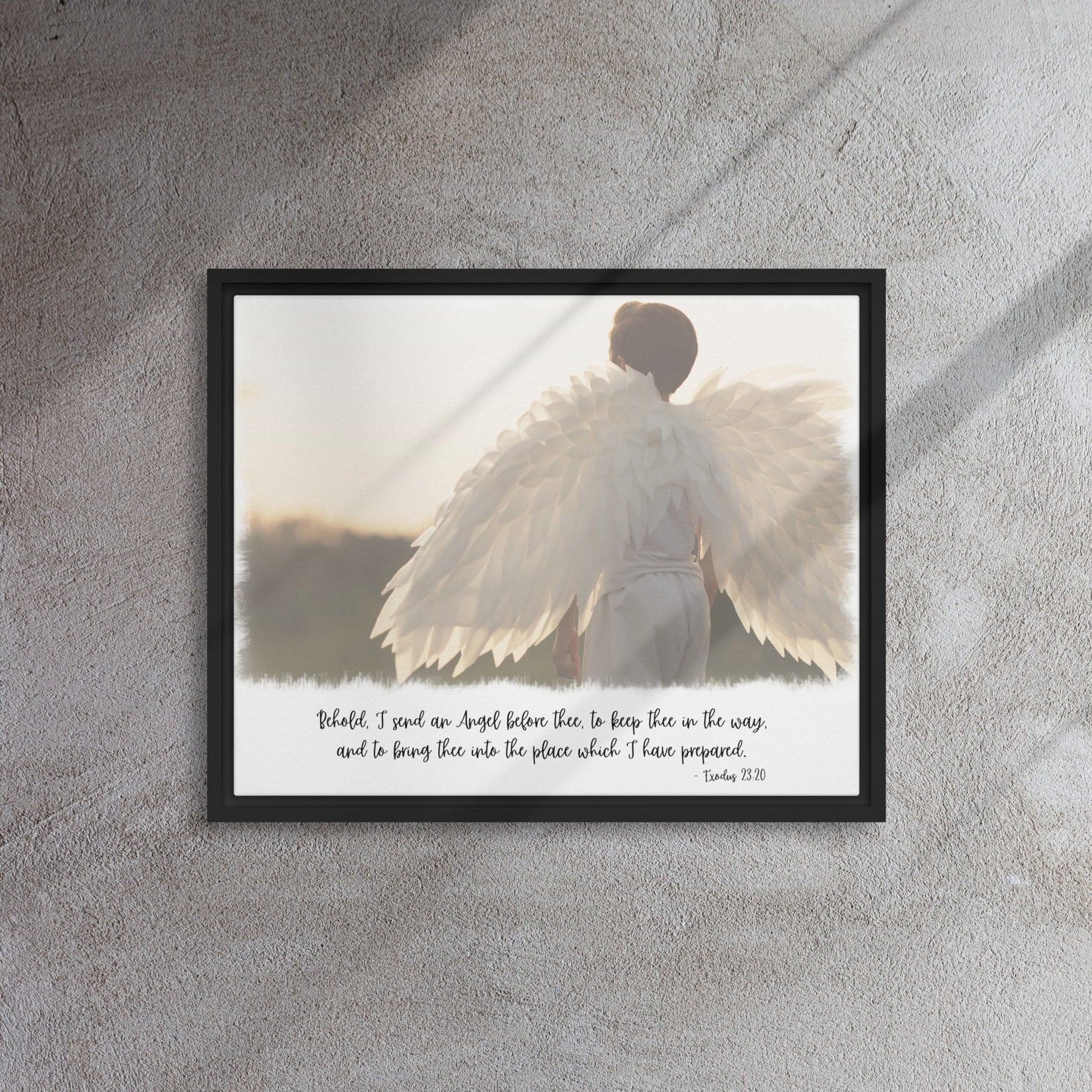 An image of the back of a realistic looking angel with spread wings looking off into the distance, with scripture below it in a script font, framed with a black frame, hangs on a gray textured wall with sunlight streaming in across it from the right.