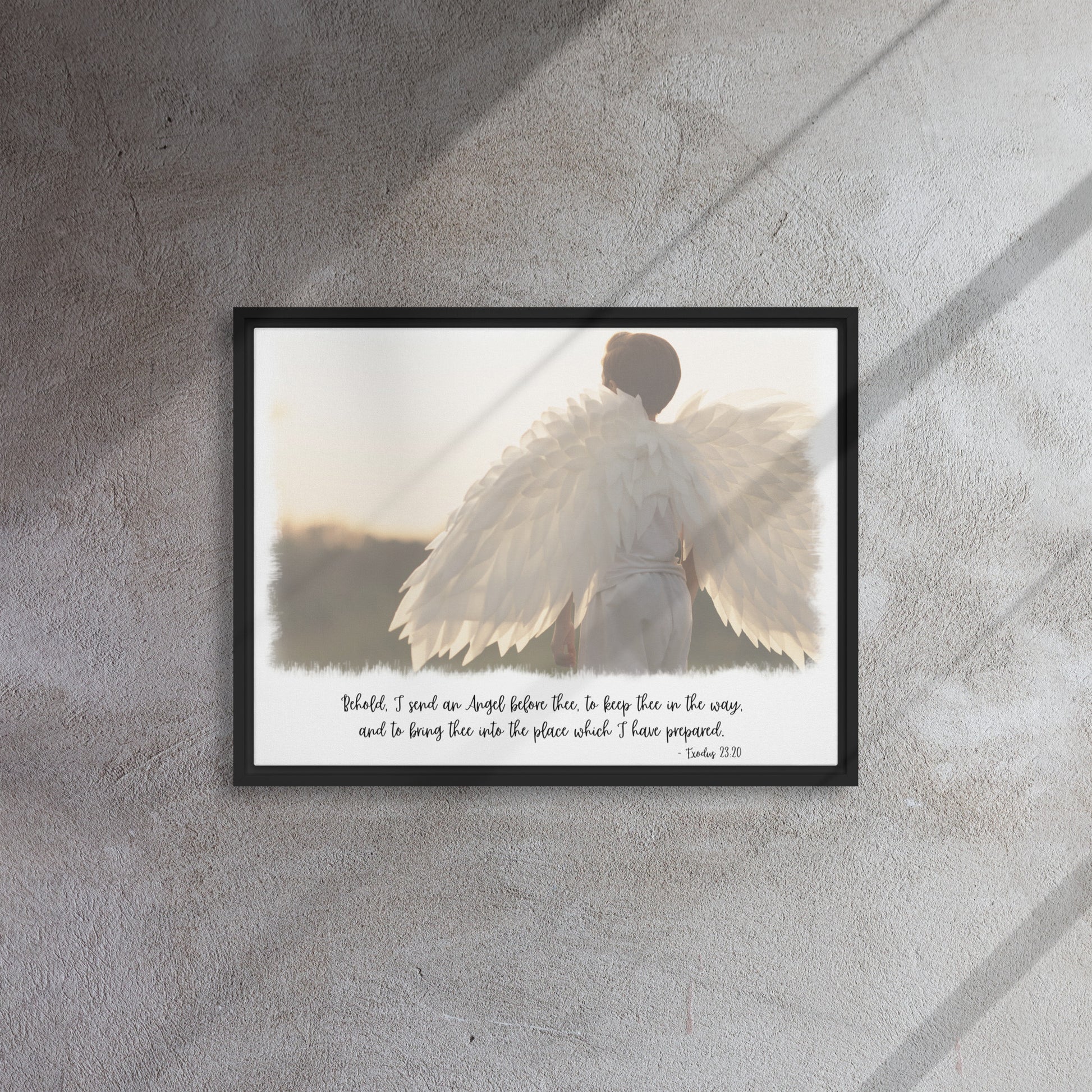 An image of the back of a realistic looking angel with spread wings looking off into the distance, with scripture below it in a script font, framed with a black frame, hangs on a gray textured wall with sunlight streaming in across it from the right.