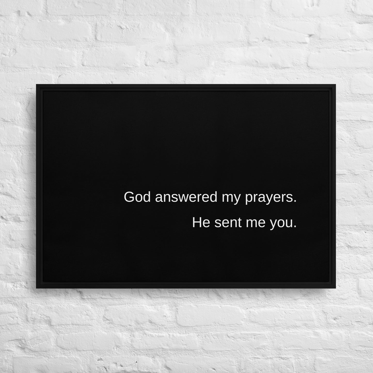 Large horizontal black stretched canvas with a black frame and white text on the canvas says:  God answered my prayers. He sent me you. And it is hanging on a white brick wall.