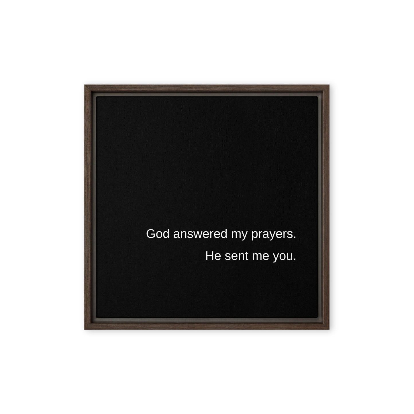 Square black stretched canvas with a brown frame on a white wall. On the black canvas, in white letters, it says:  God answered my prayers. He sent me you.