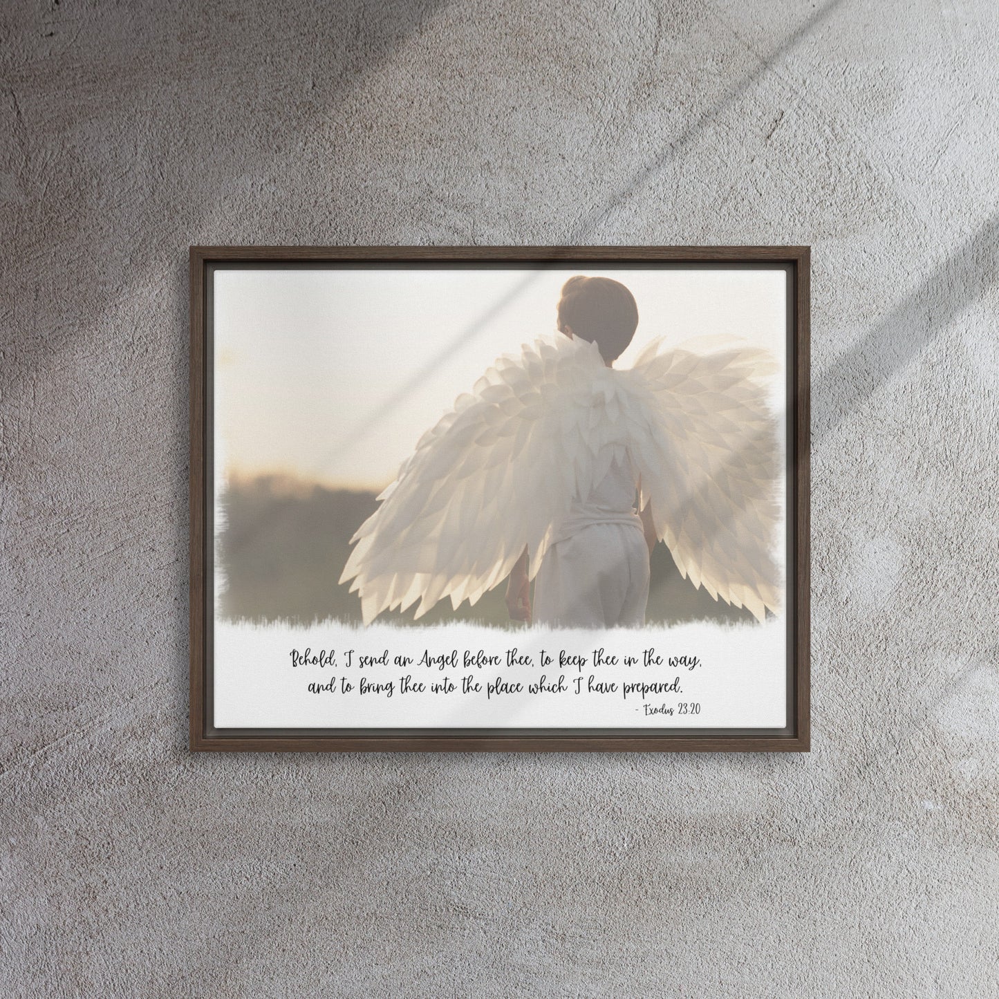 An image of the back of a realistic looking angel with spread wings looking off into the distance, with scripture below it in a script font, framed with a brown frame, hangs on a gray textured wall with sunlight streaming in across it from the right.