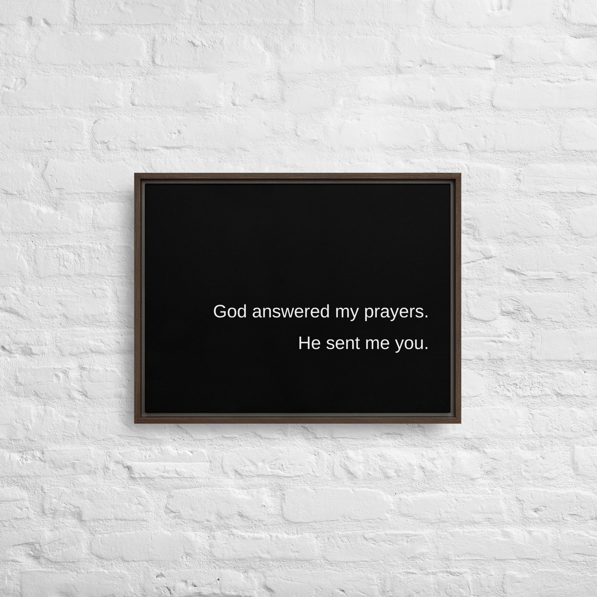 Horizontal black stretched canvas with white text on it, with a brown wooden frame, hanging on a white brick wall. The canvas has white words on it that say:  God answered my prayers. He sent me you.