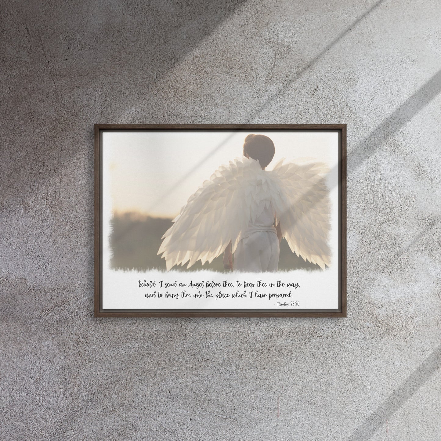 An image of the back of a realistic looking angel with spread wings looking off into the distance, with scripture below it in a script font, framed with a brown frame, hangs on a gray textured wall with sunlight streaming in across it from the right.