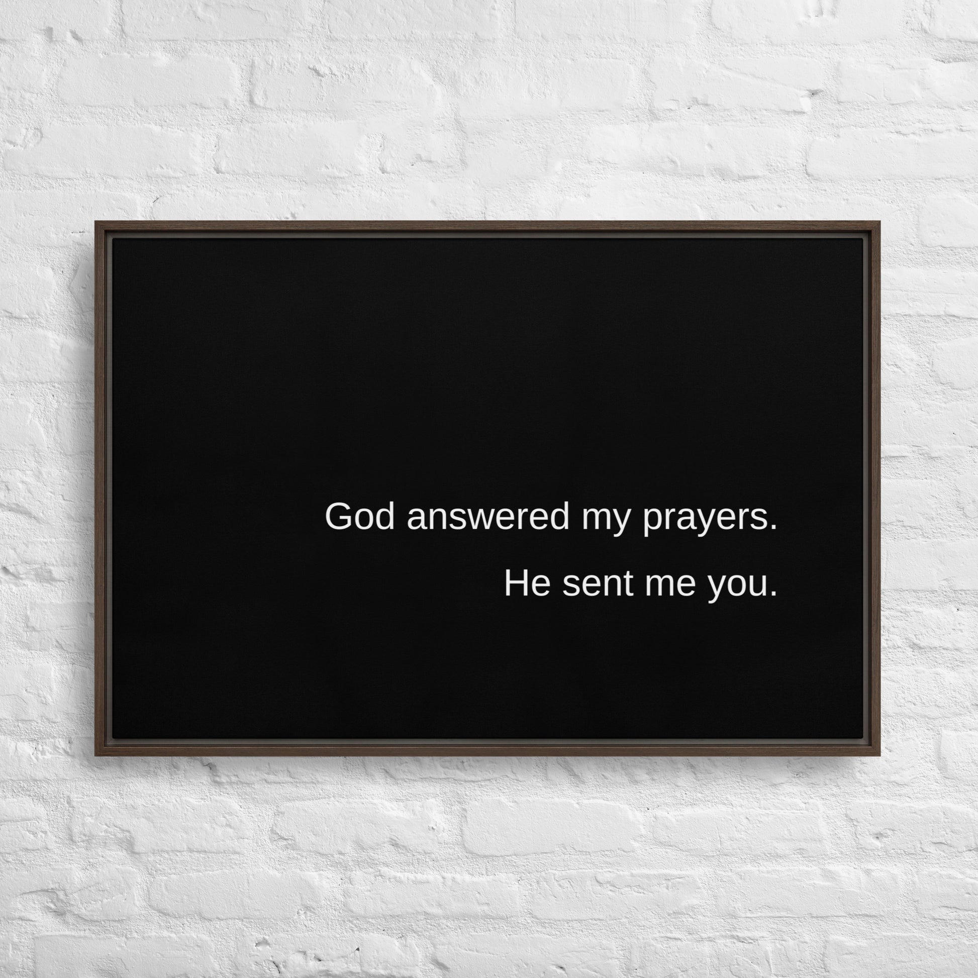 Horizontal framed art piece on a white brick wall. The art is solid black with a brown frame and the art itself is a solid black canvas with white words that say:  God answered my prayers. He sent me you.