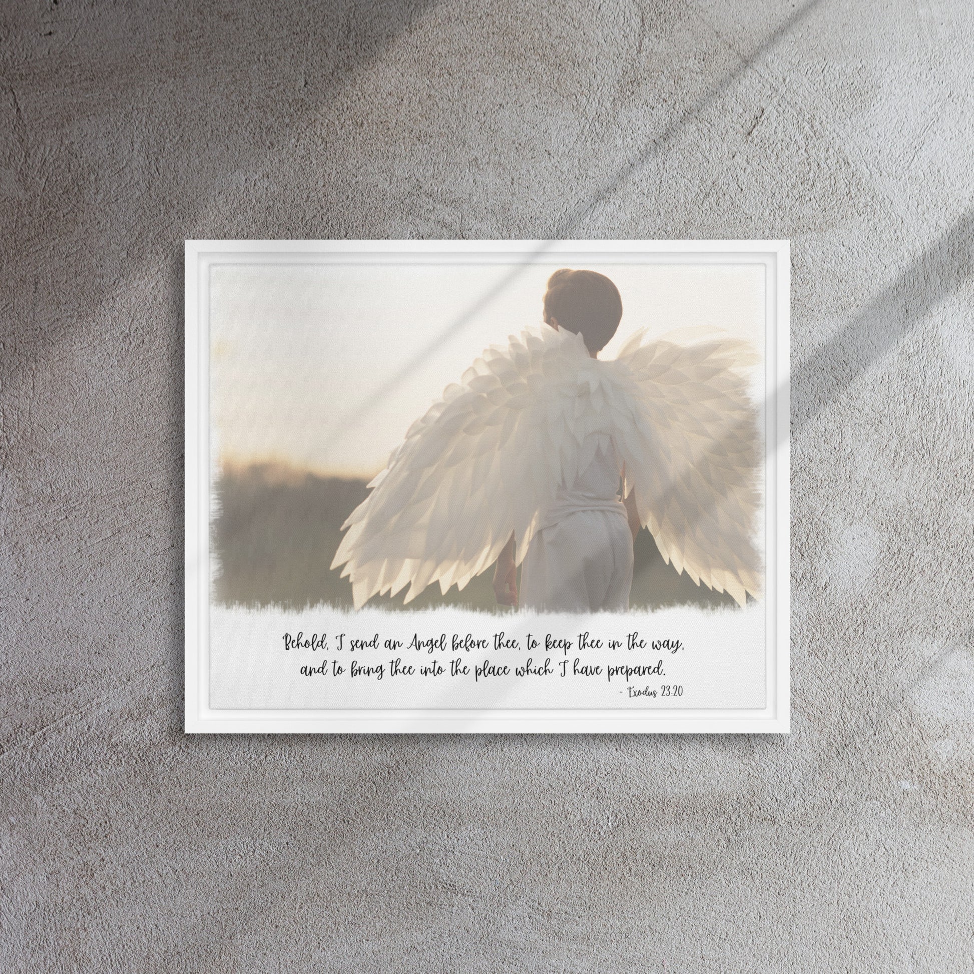 An image of the back of a realistic looking angel with spread wings looking off into the distance, with scripture below it in a script font, framed with a white frame, hangs on a gray textured wall with sunlight streaming in across it from the right.