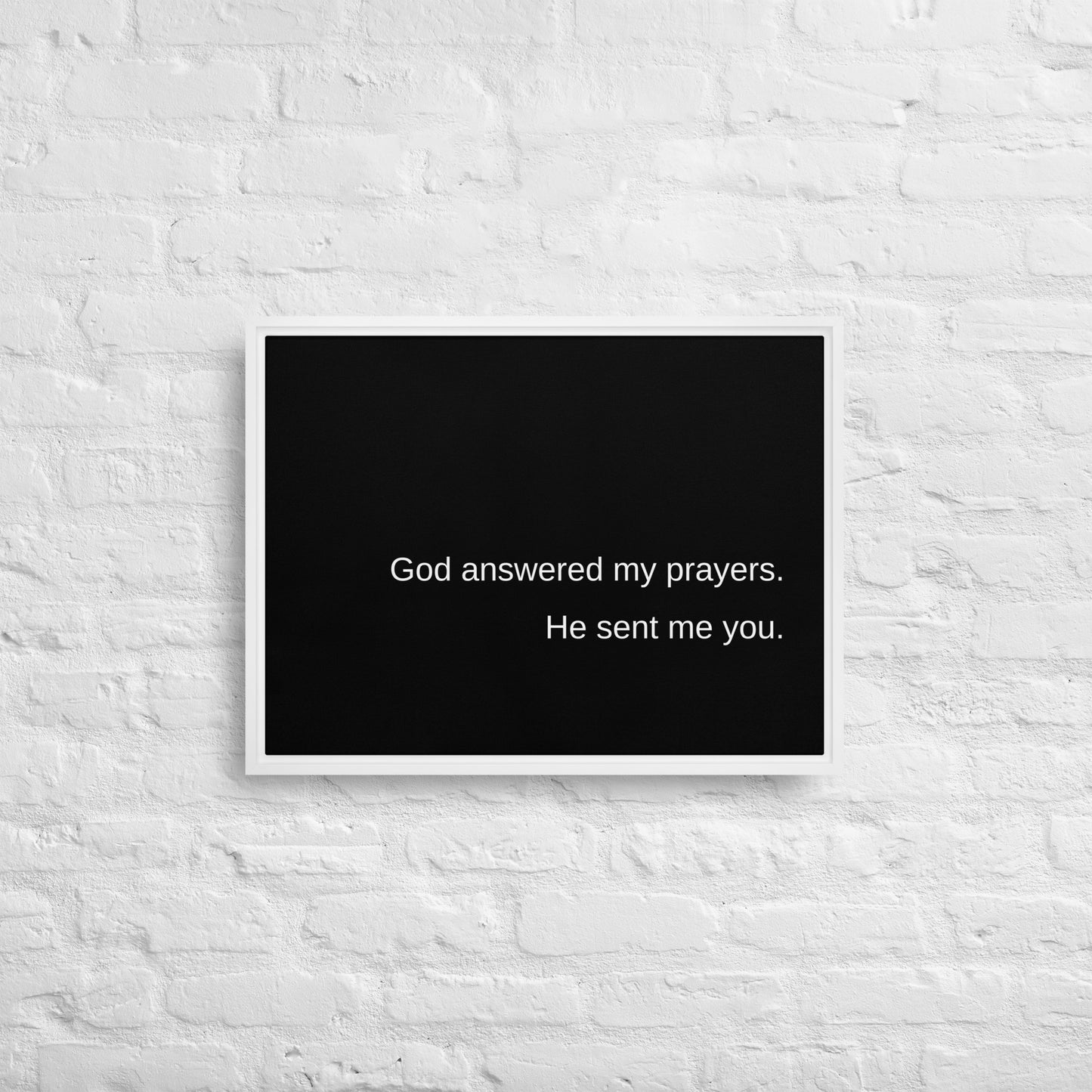 Horizonal art piece with a white frame and white words on a black background hanging on a white brick wall. The words on the canvas say:  God answered my prayers. He sent me you.